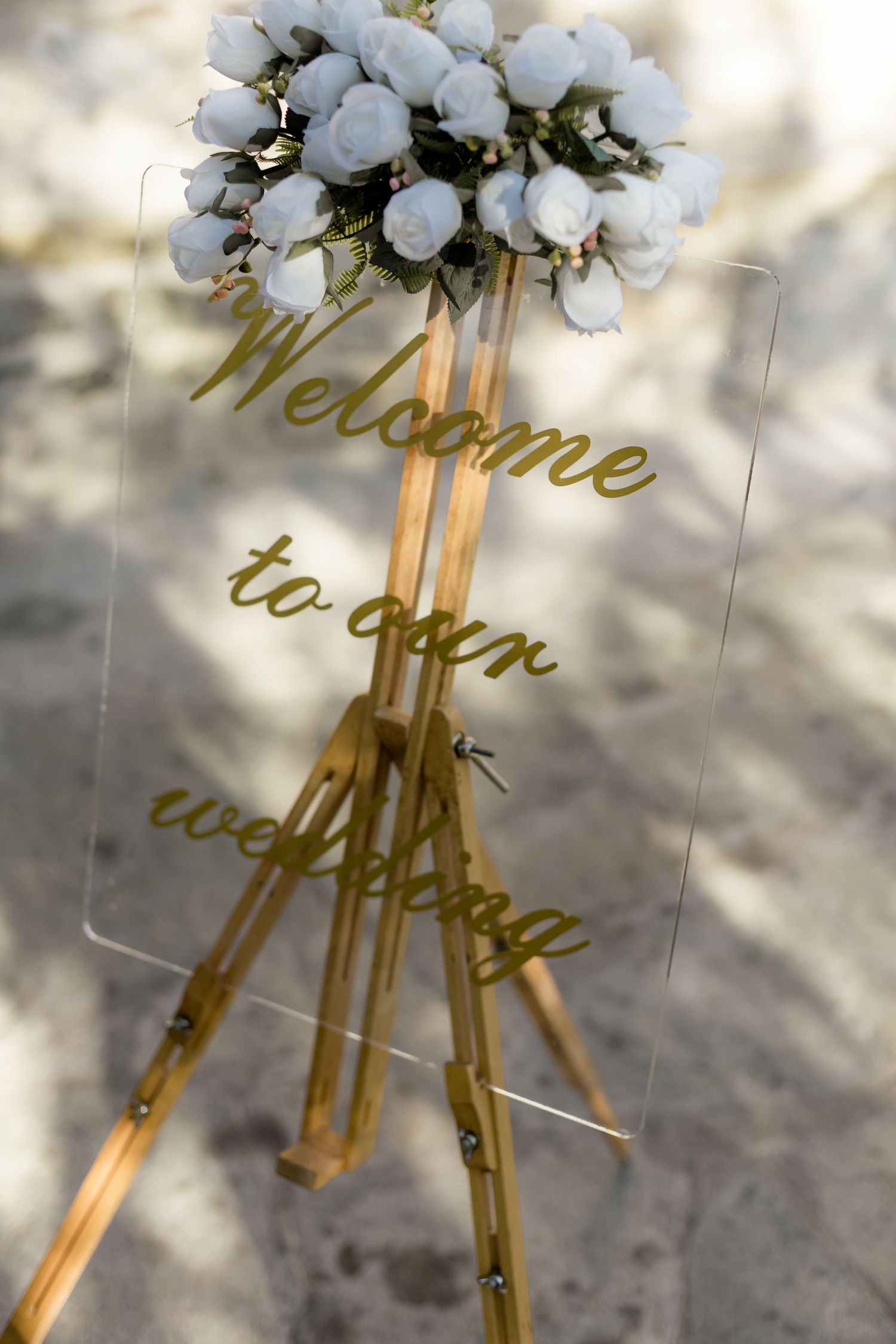 Welcome sign decorated with white flowers and gold lettering on clear acrylic stand.