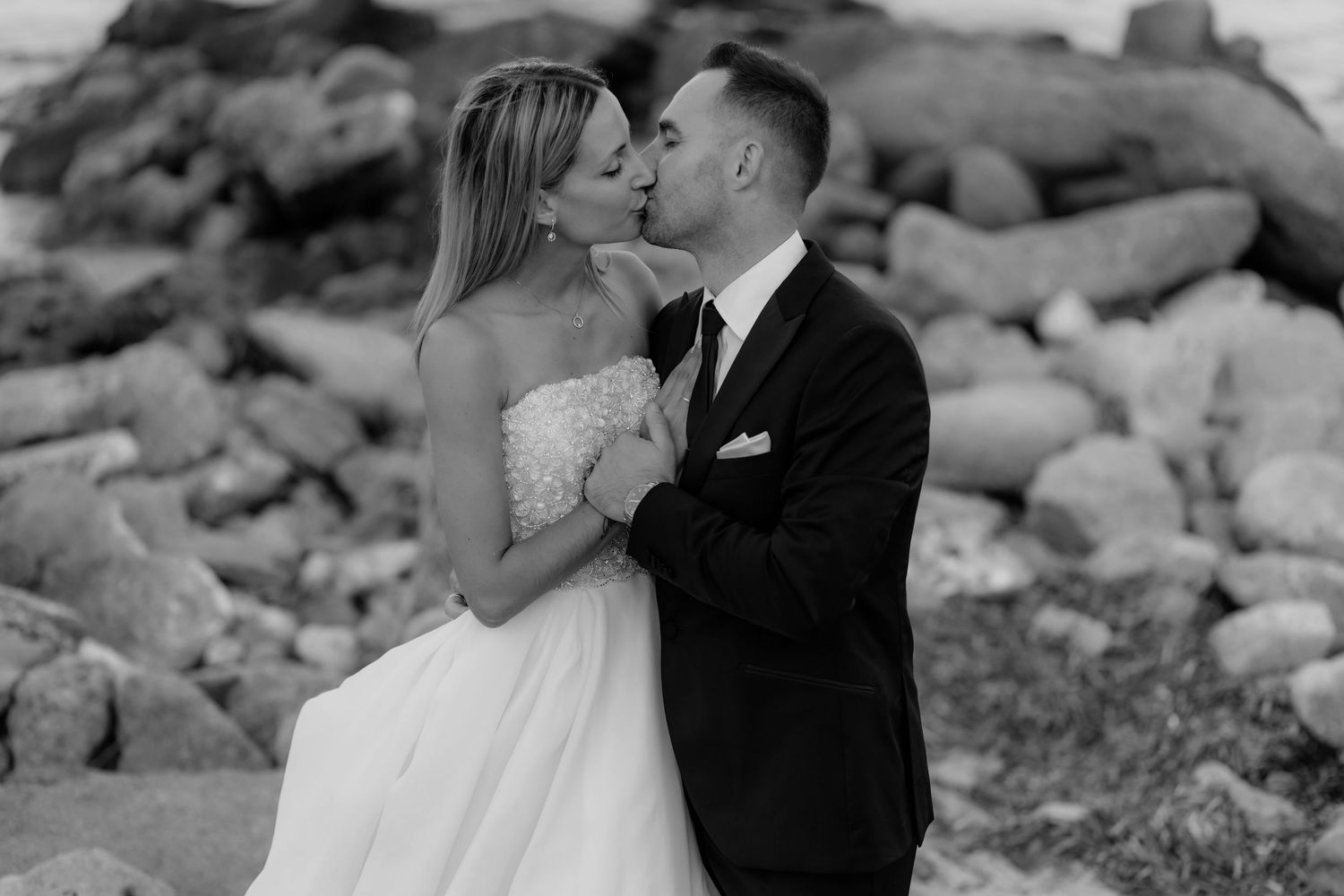 Black and white portrait of newlyweds sharing an intimate moment among coastal rocks.