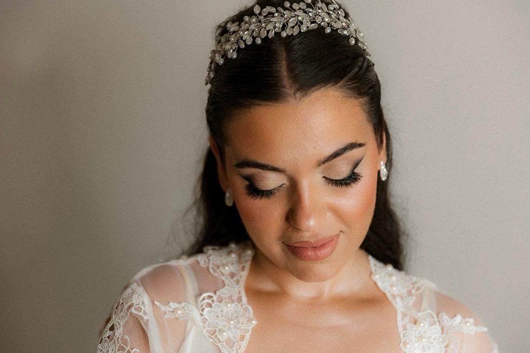 Bride wearing a crystal tiara and lace wedding dress with elegant makeup and long lashes, looking down gracefully.