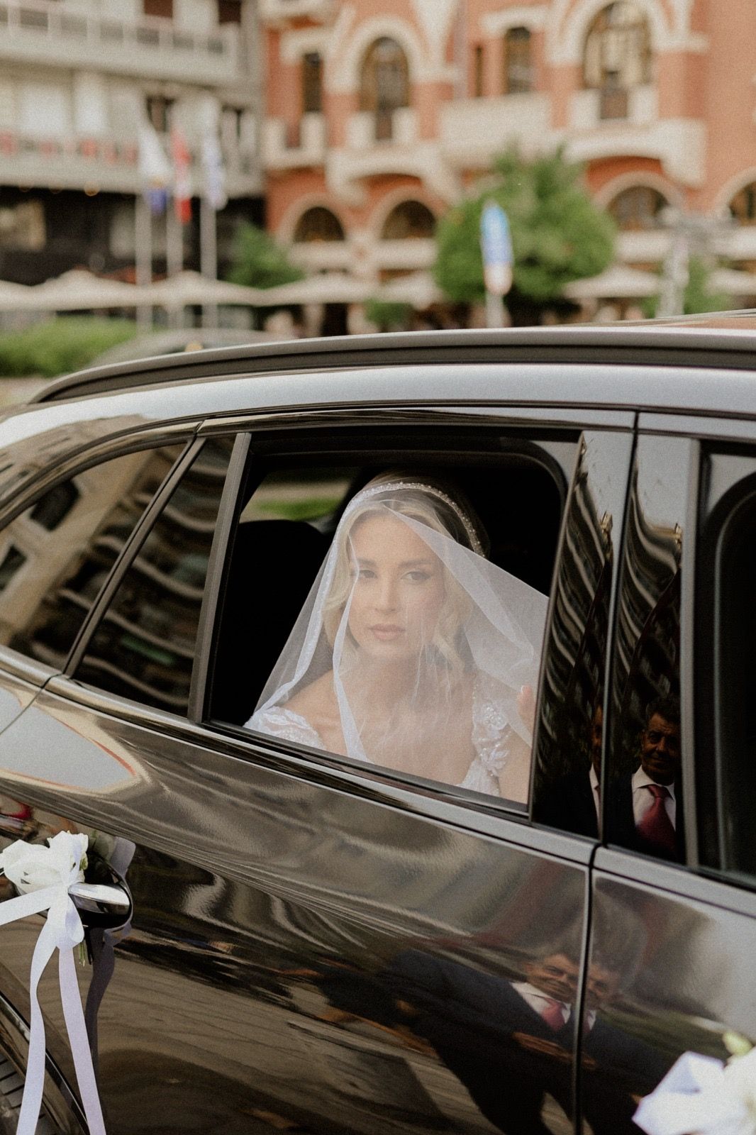 Bride in white veil sitting inside luxury black car with decorative bows.