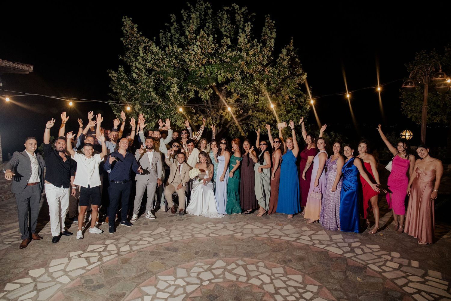 Large wedding party celebrates on an outdoor patio at night with raised arms.