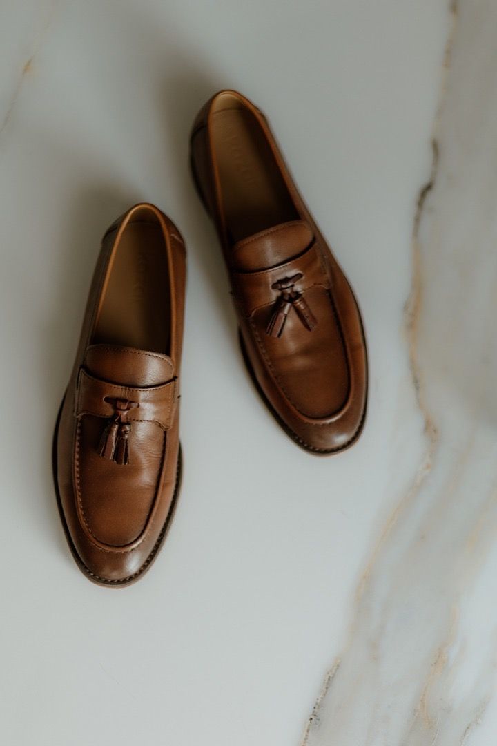 Brown leather tassel loafers displayed on white marble surface.