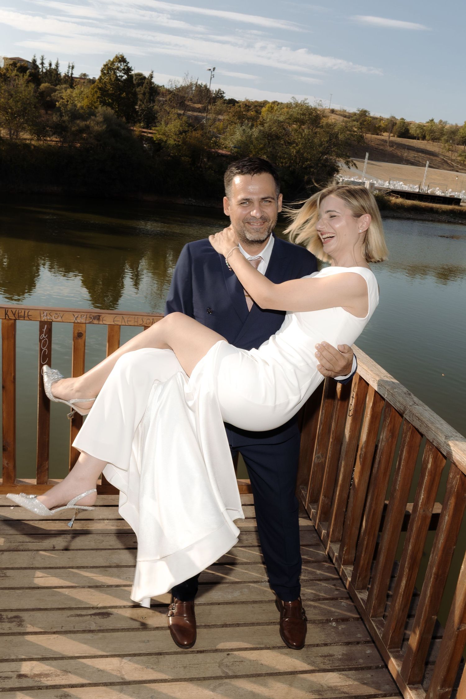 Couple on wooden dock by lake, with groom carrying bride in romantic pose.