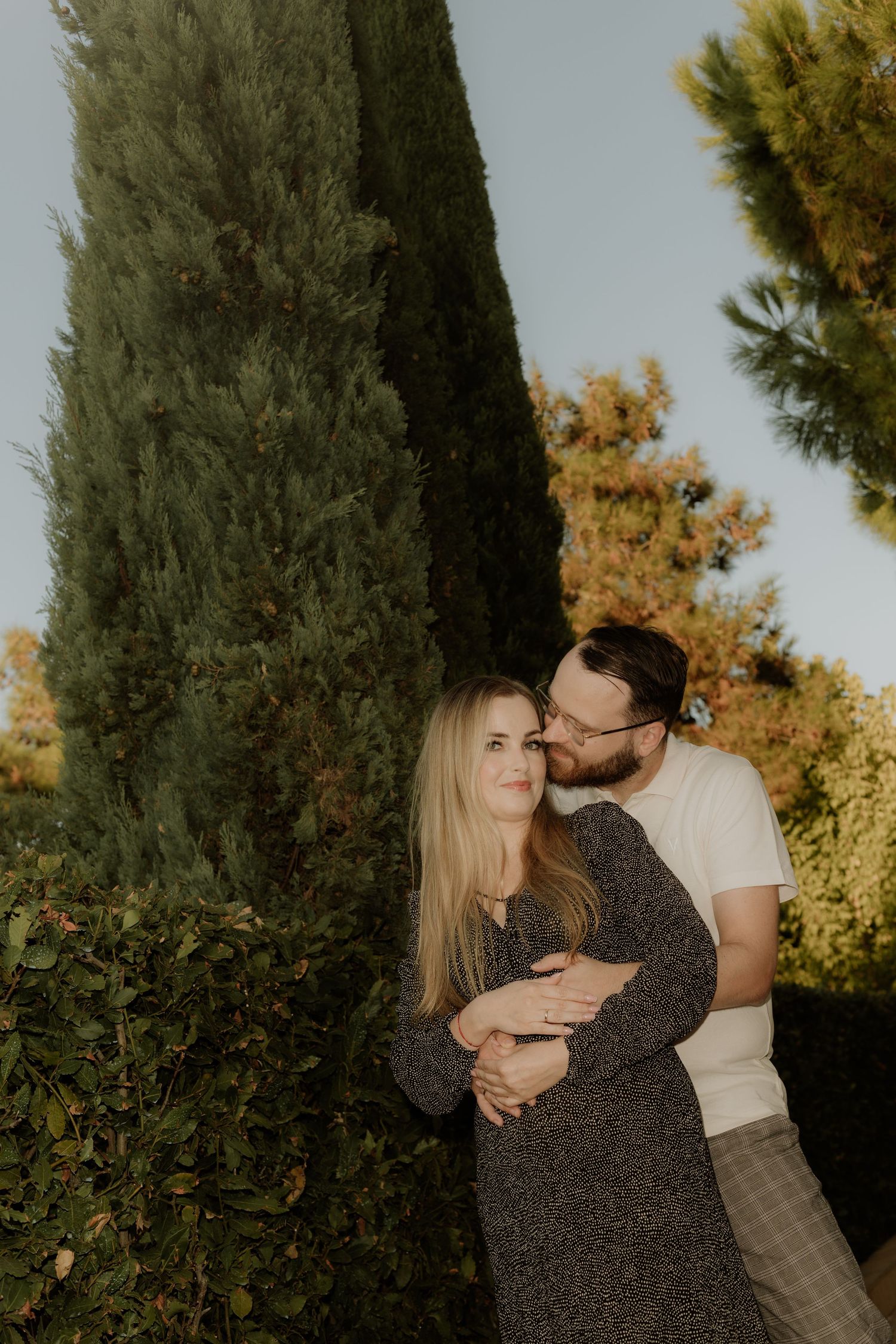 Romantic outdoor portrait of a couple embracing near cypress trees in warm evening light.