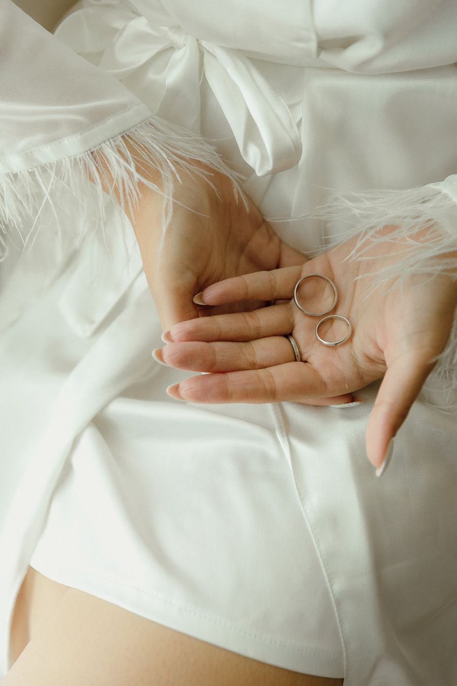 Bride in white feathered robe holding two gold wedding rings in her palm, showcasing elegant bridal jewelry.