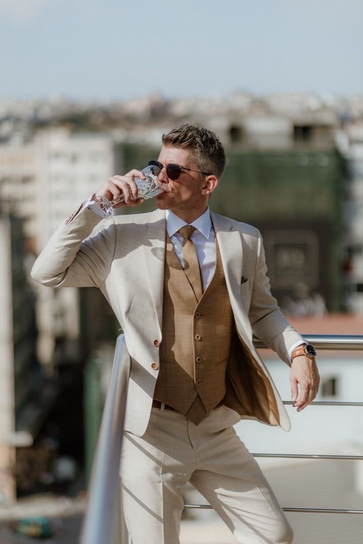 Stylish groom in cream suit and brown vest drinking from glass on sunny terrace.