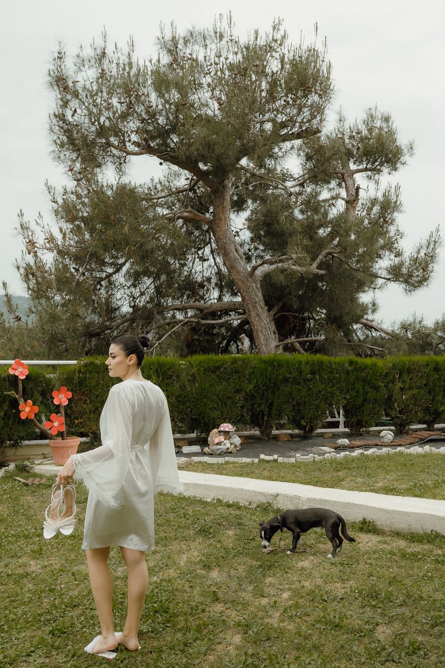 Woman in white robe holding shoes stands in garden with black dog and large pine tree backdrop.
