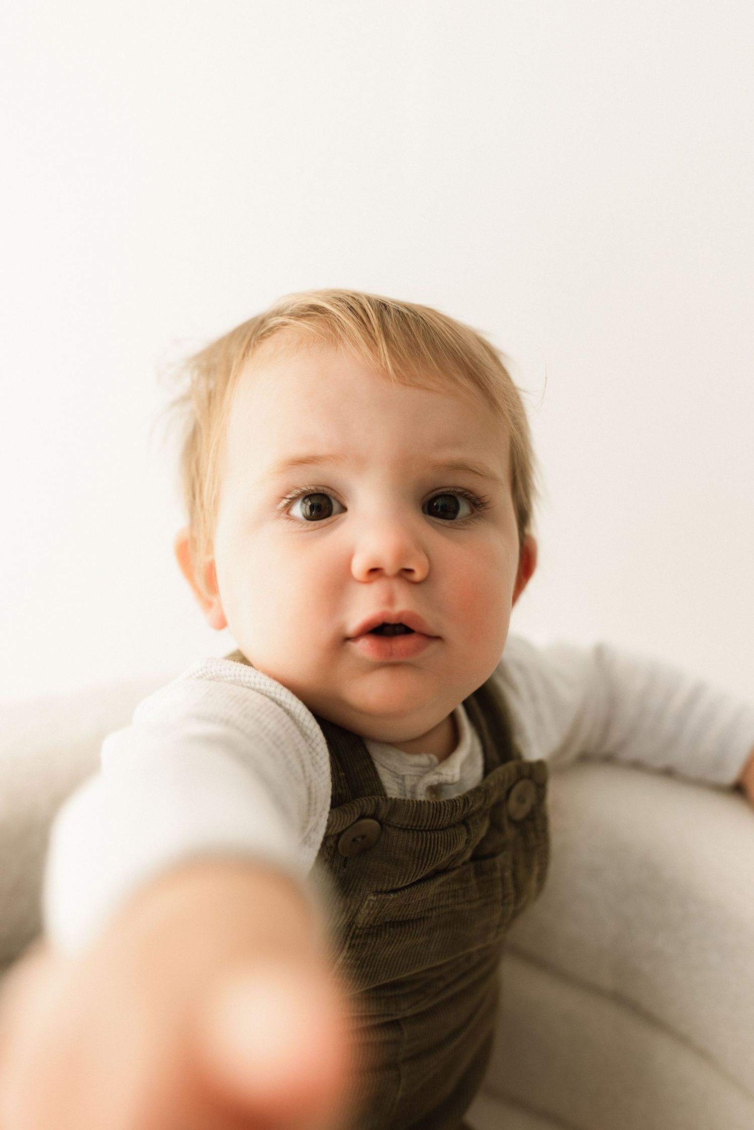 A young child in brown overalls and white shirt lays on a white surface looking upward with a gentle expression.