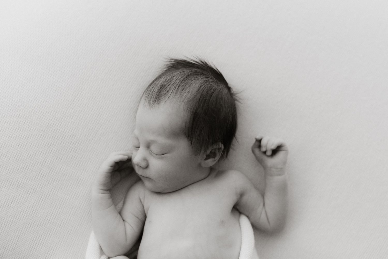 Black and white newborn photo of a sleeping baby with dark hair, tiny raised hand, on white background.