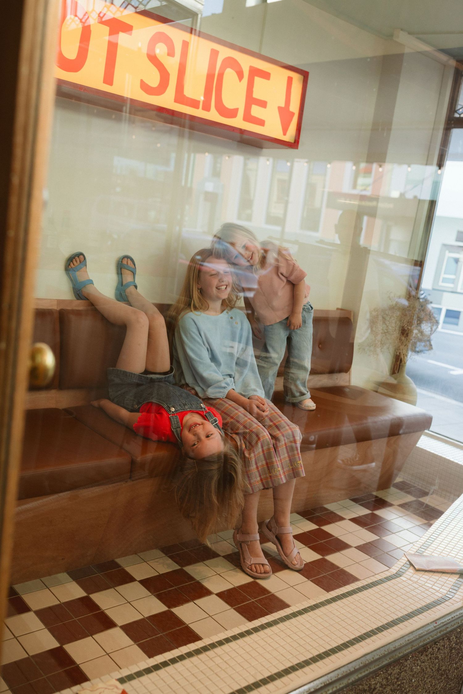 Two children sit in a pizza shop window beneath a glowing red Hot Slice sign with checkered tile floors.
