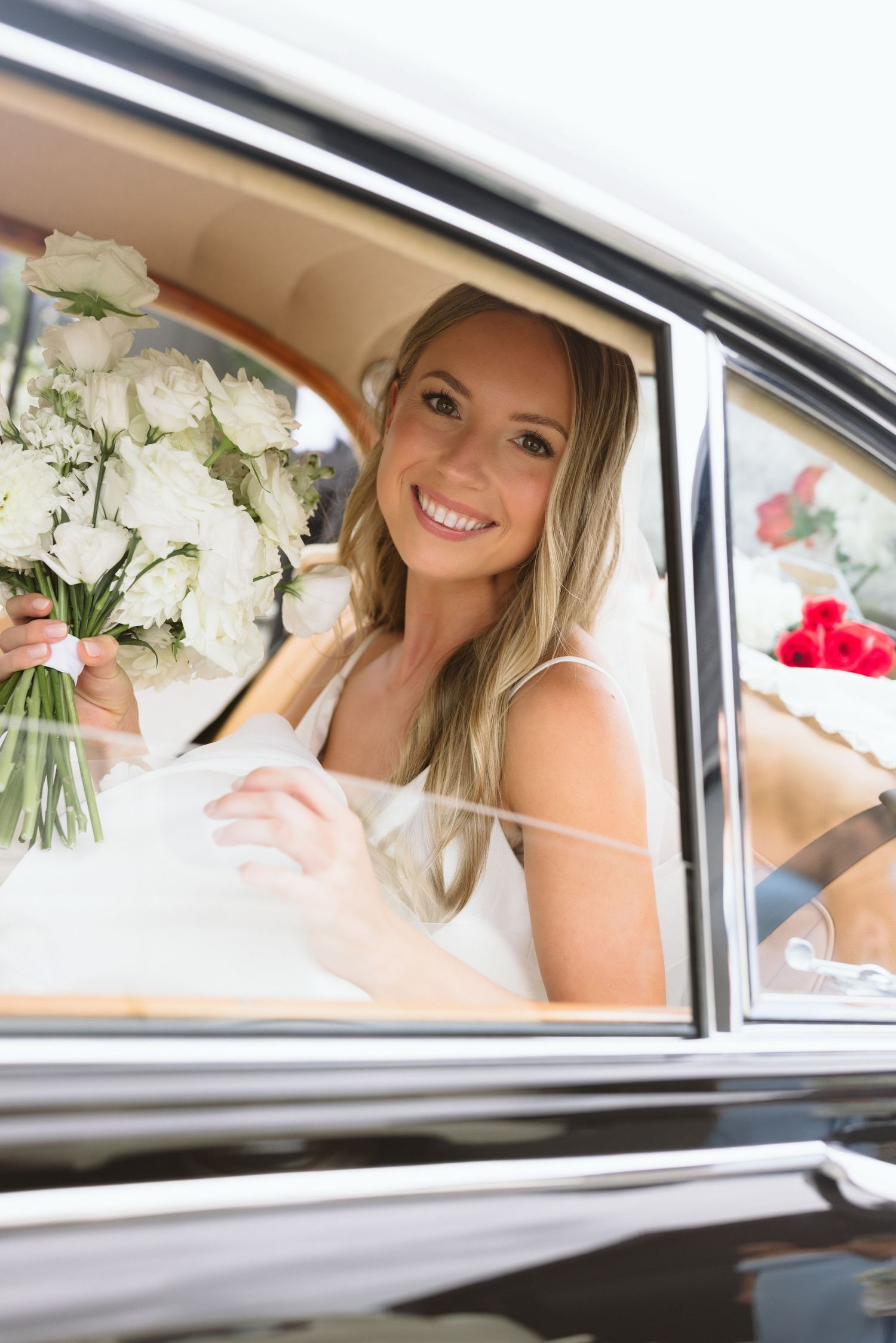 A bride sits in a classic car with white floral bouquets visible through the window.