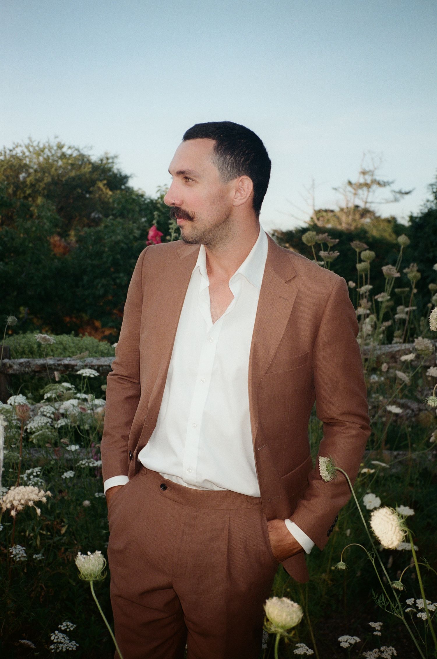 A person in a brown suit and white shirt stands in a garden with blooming flowers during golden hour.