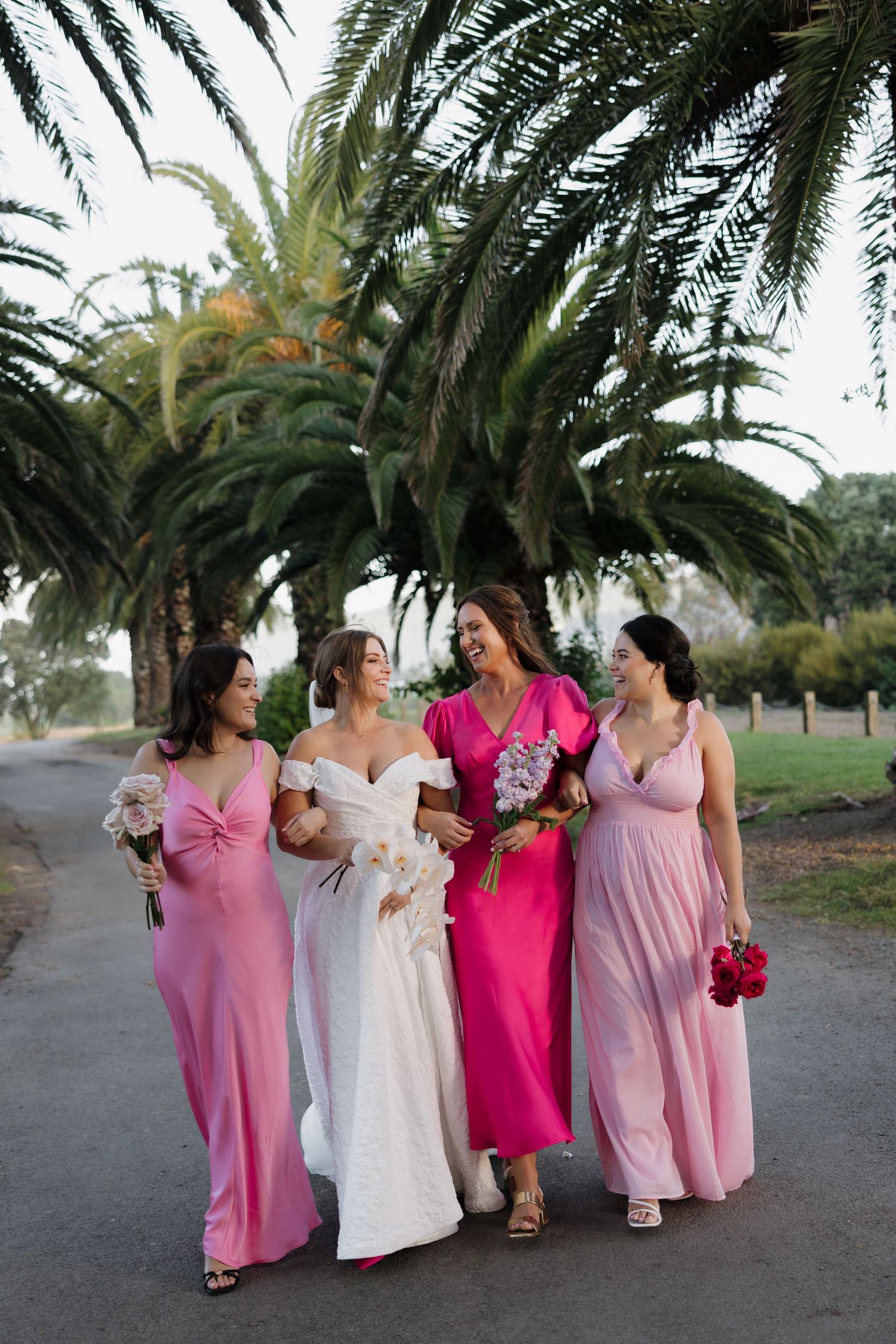 Wedding party in pink and white dresses walking beneath tropical palm trees.