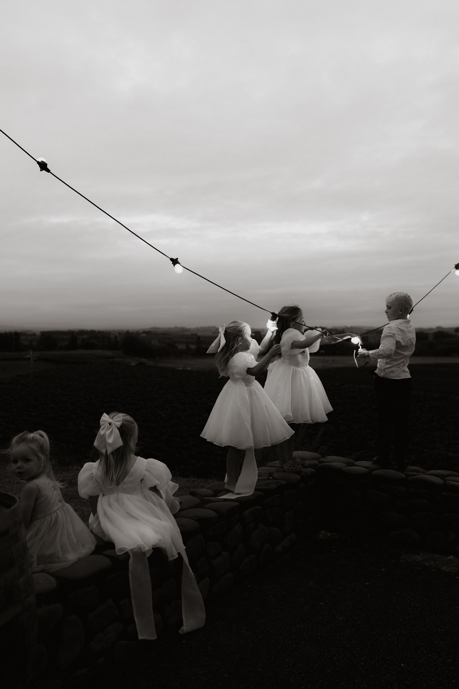 Black and white photo of flower girls in white dresses and a boy hanging string lights on a stone wall at dusk.