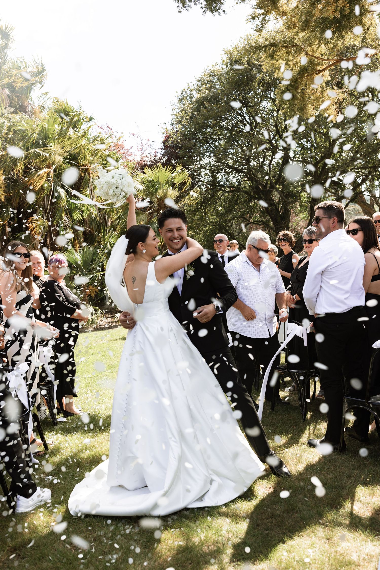 Wedding celebration with guests throwing white confetti in outdoor garden setting.