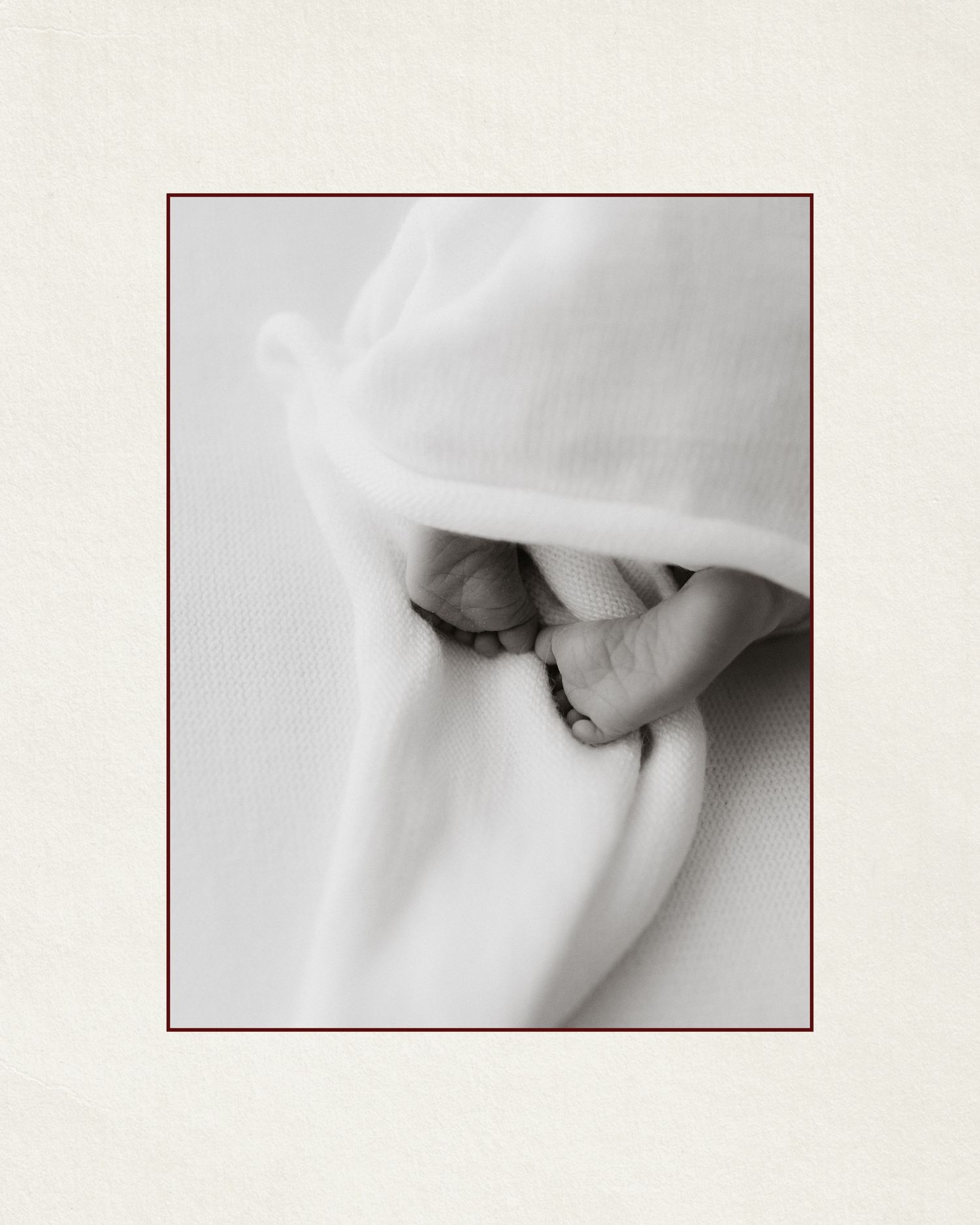 Black and white photo of a newborn baby wrapped in white fabric, peacefully sleeping with tiny hands visible.