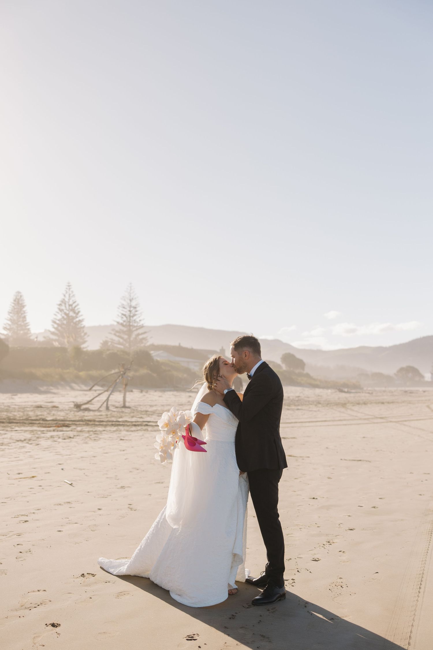 Romantic beach wedding photo with couple silhouetted against mountains and pine trees.