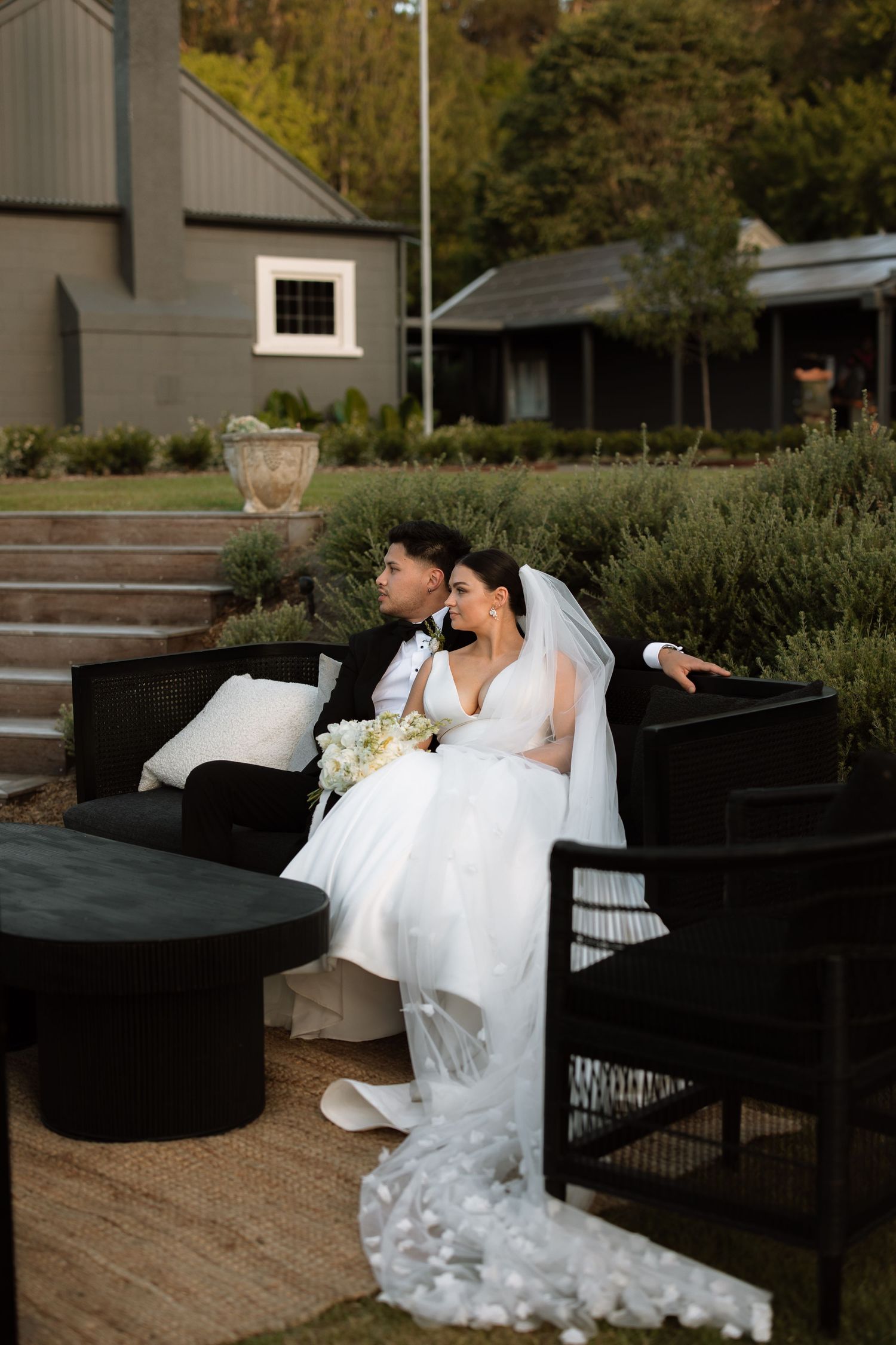 Bride relaxes on outdoor furniture in elegant white wedding gown.