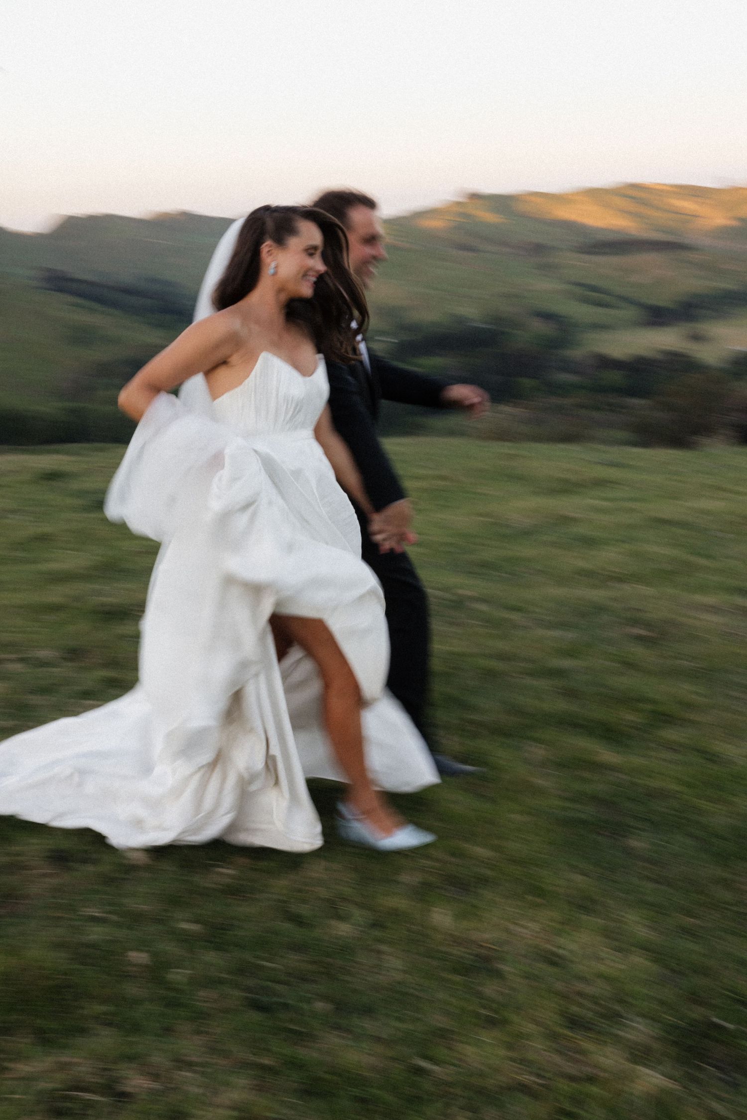 A person in a flowing white dress runs and spins through a grassy hillside at sunset.