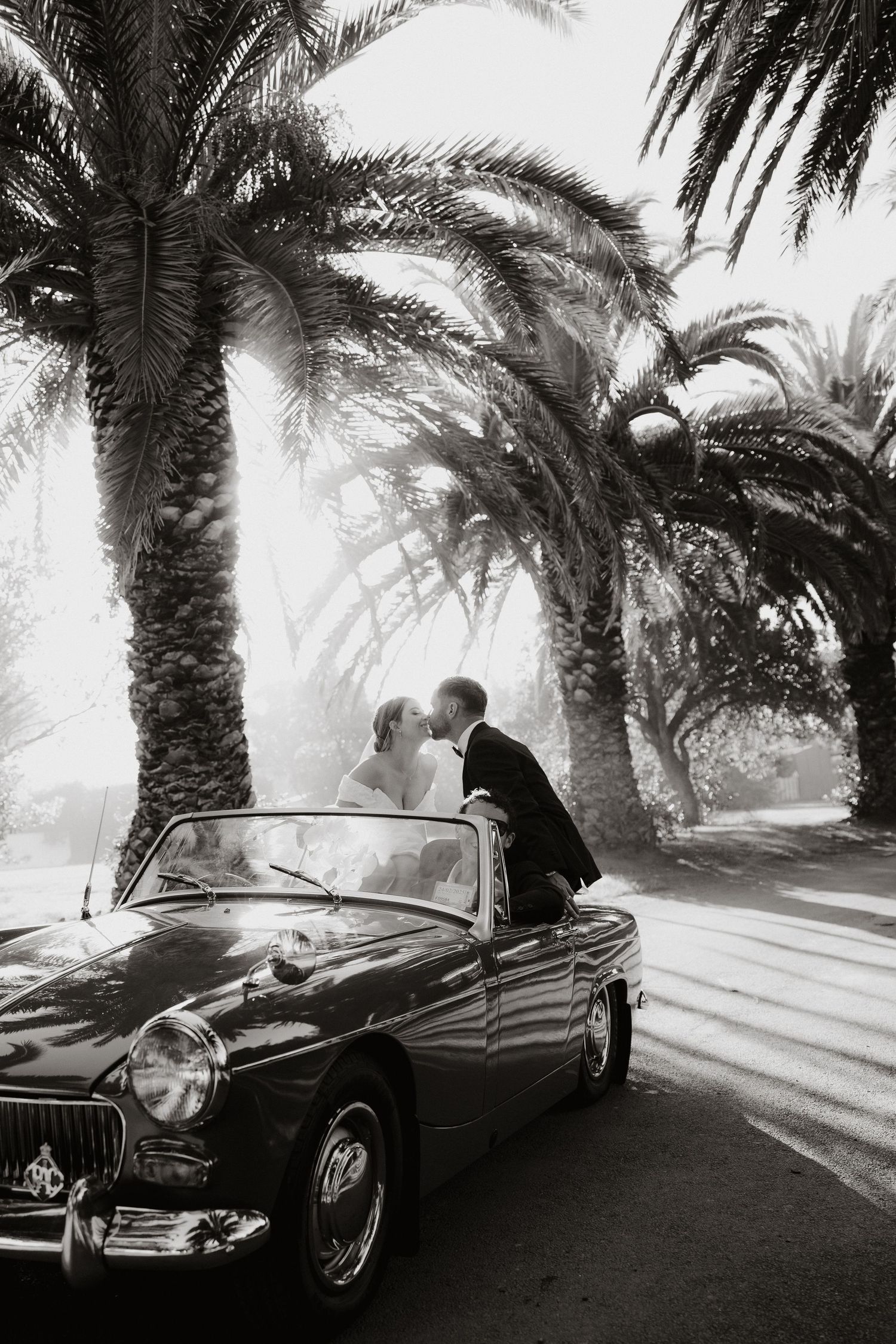 Romantic black and white photo of couple with classic car under palm trees.