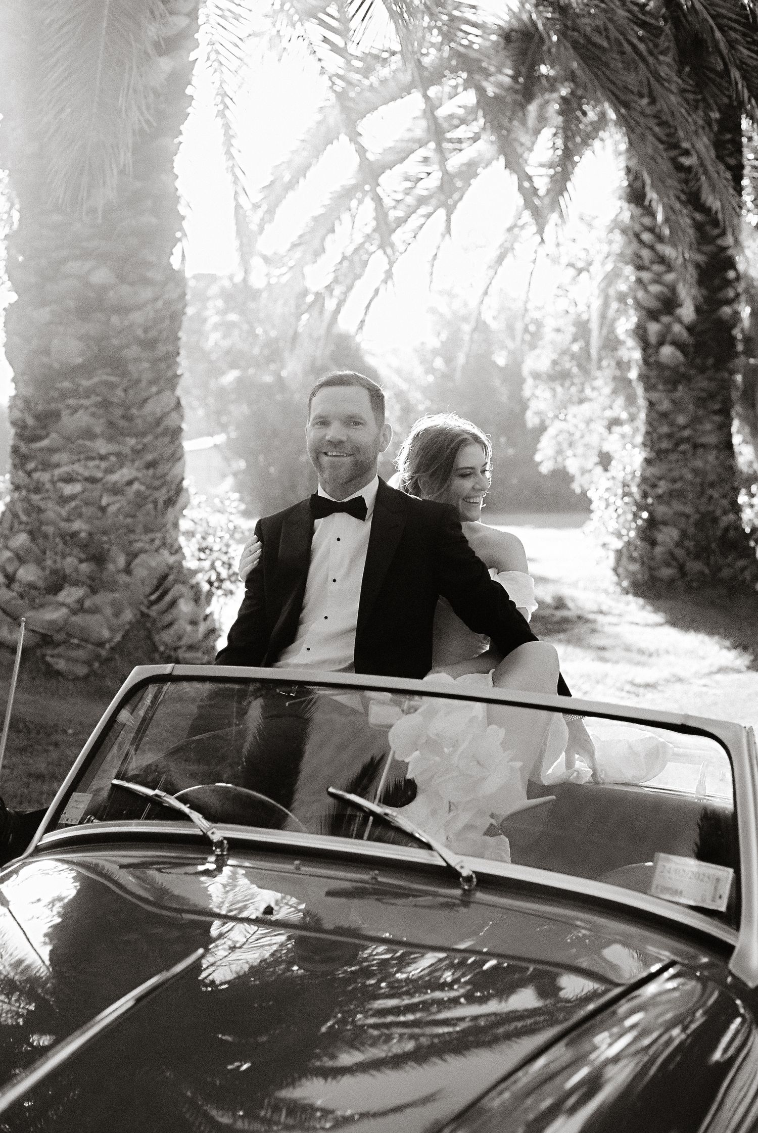 A classic black and white photo of newlyweds in a vintage convertible under palm trees.