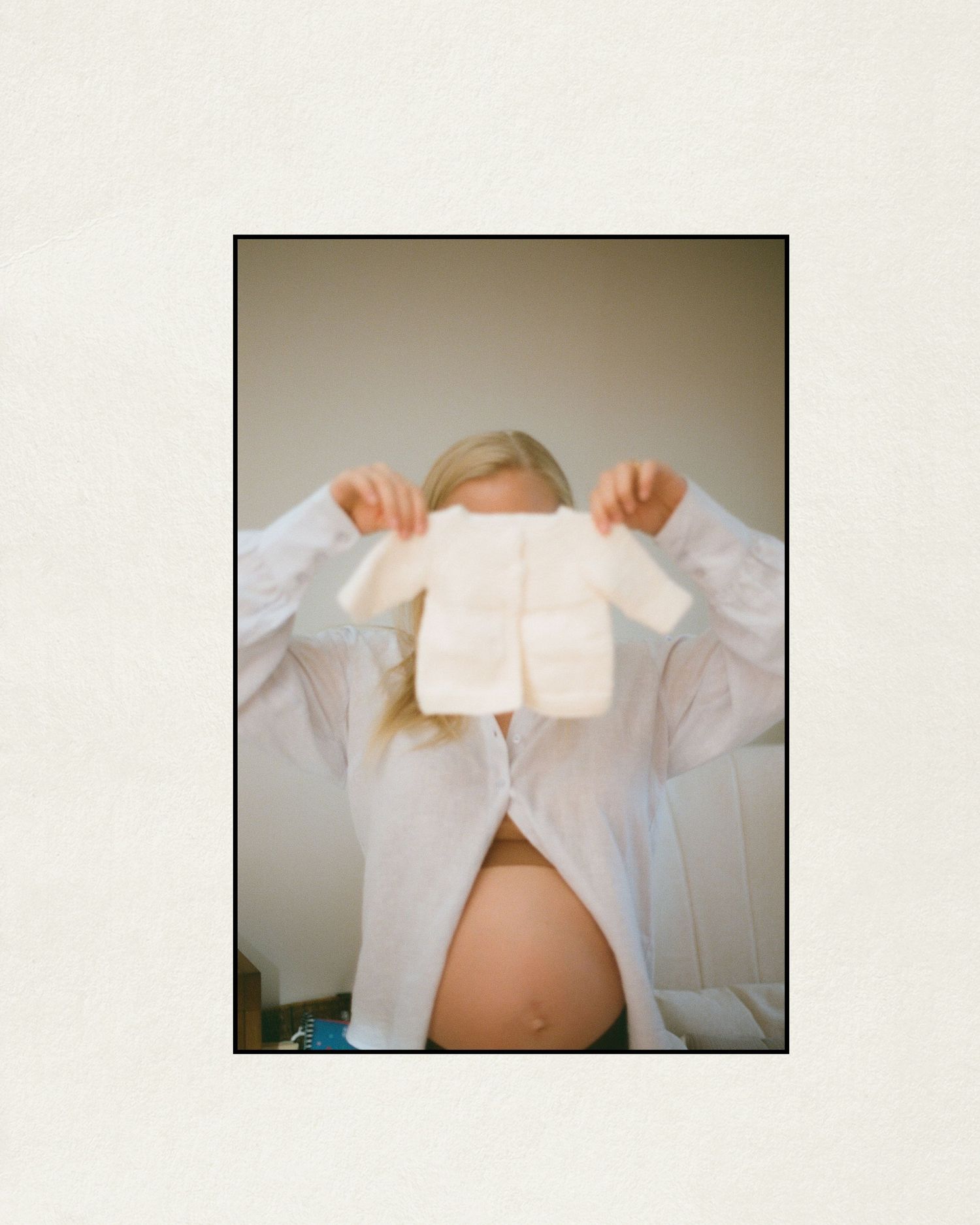 Pregnant woman in white shirt holding tiny white knit baby outfit over her face, showing bare baby bump.