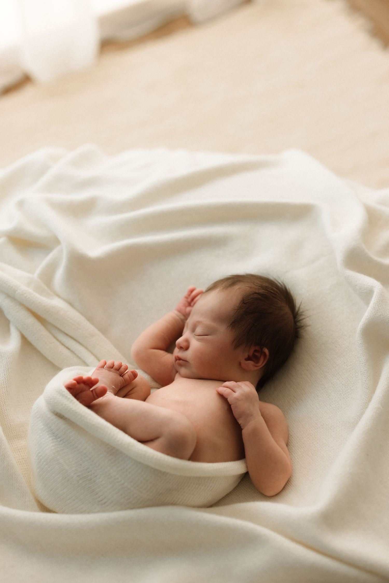 Peaceful newborn baby sleeping on a soft white blanket, with dark hair and tiny hands tucked close.
