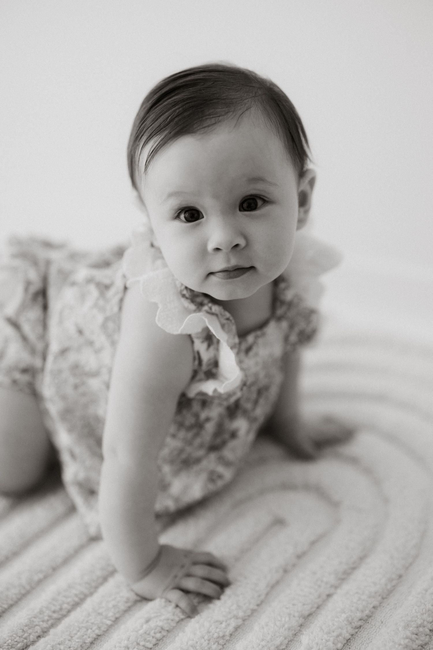 Baby in floral dress crawling on textured white surface during photo session.