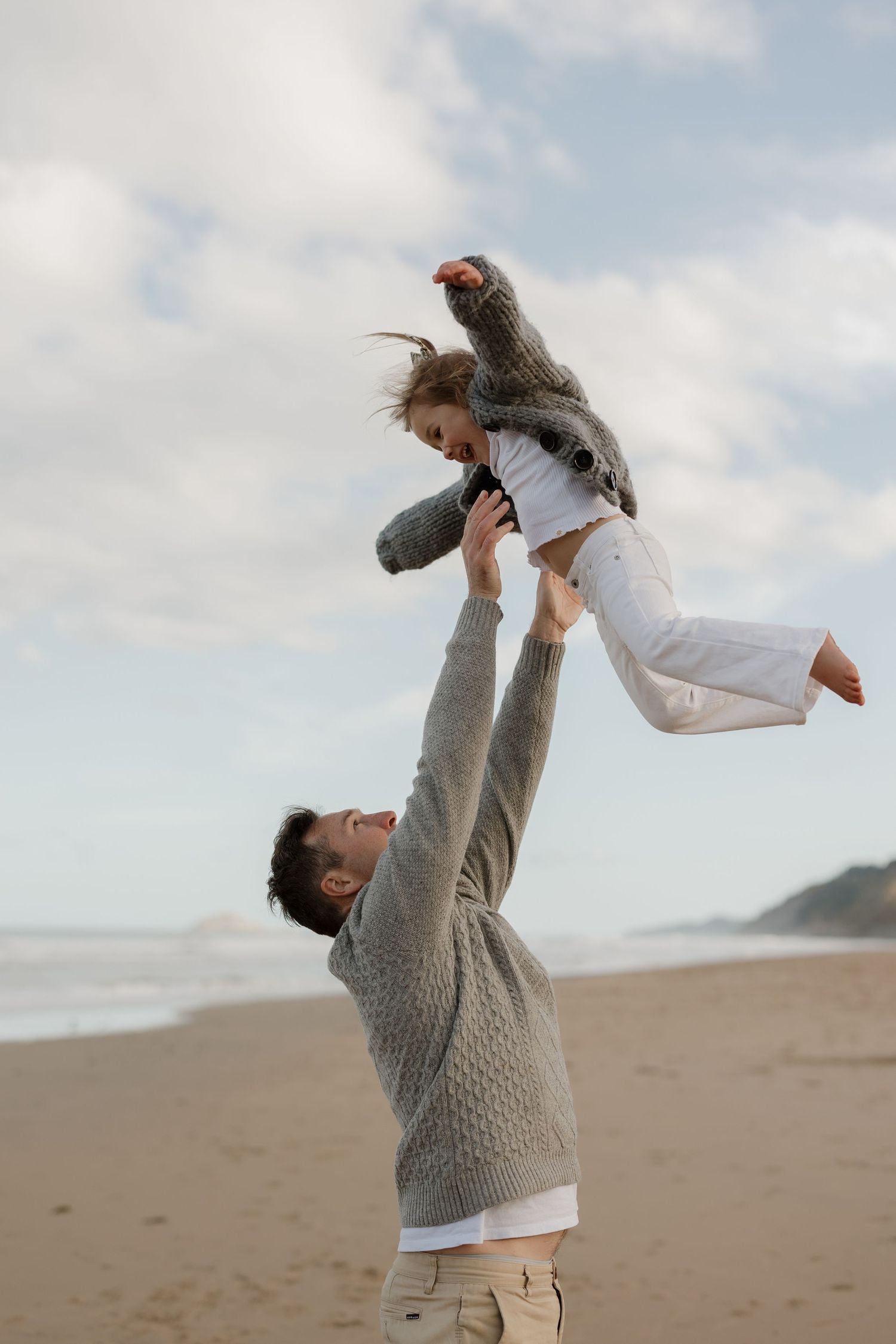 Joyful playtime moments on beach with adult lifting child in white dress against cloudy sky.