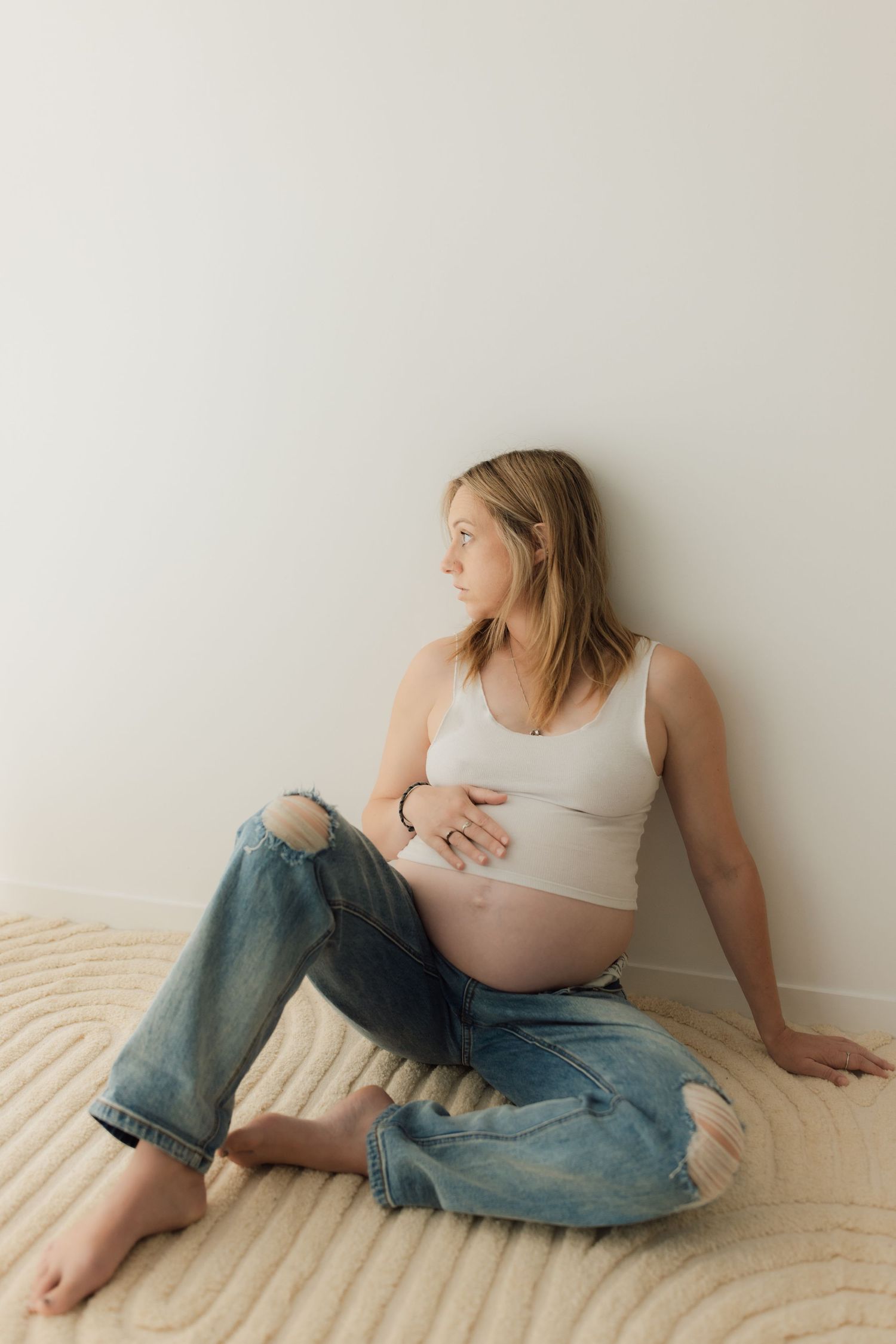 Expectant mother in casual wear sits reflectively against white wall on beige carpet.