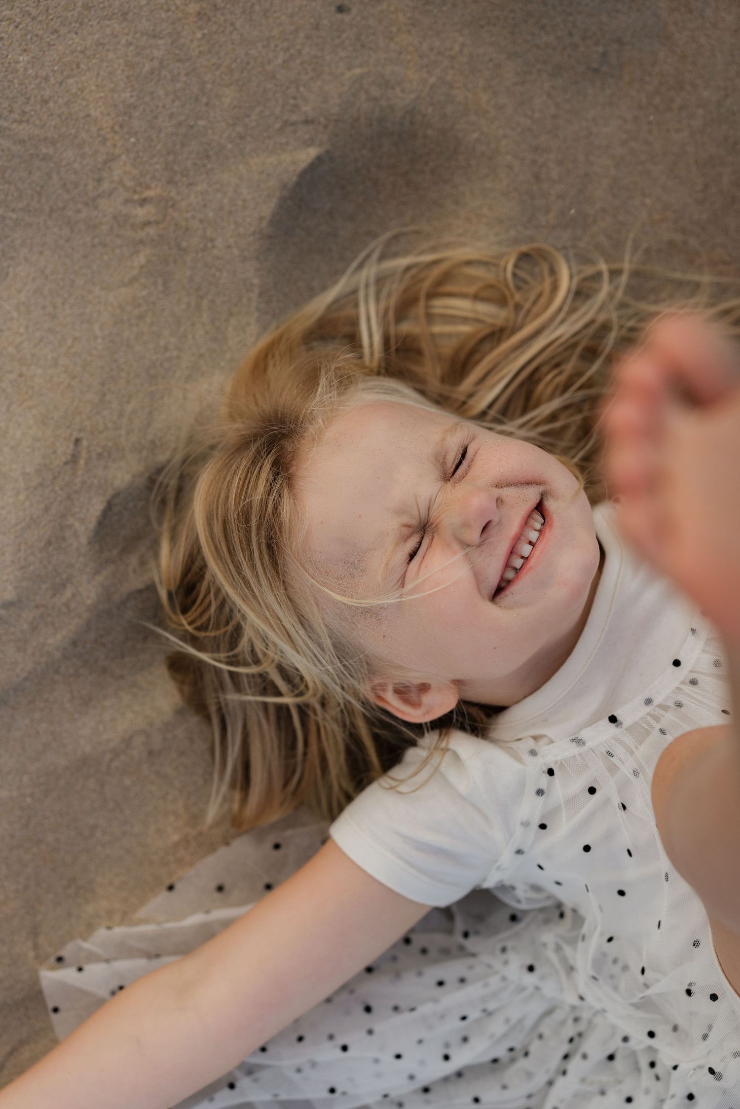 Joyful child laying on floor wearing white polka dot dress while laughing with blonde hair spread out.