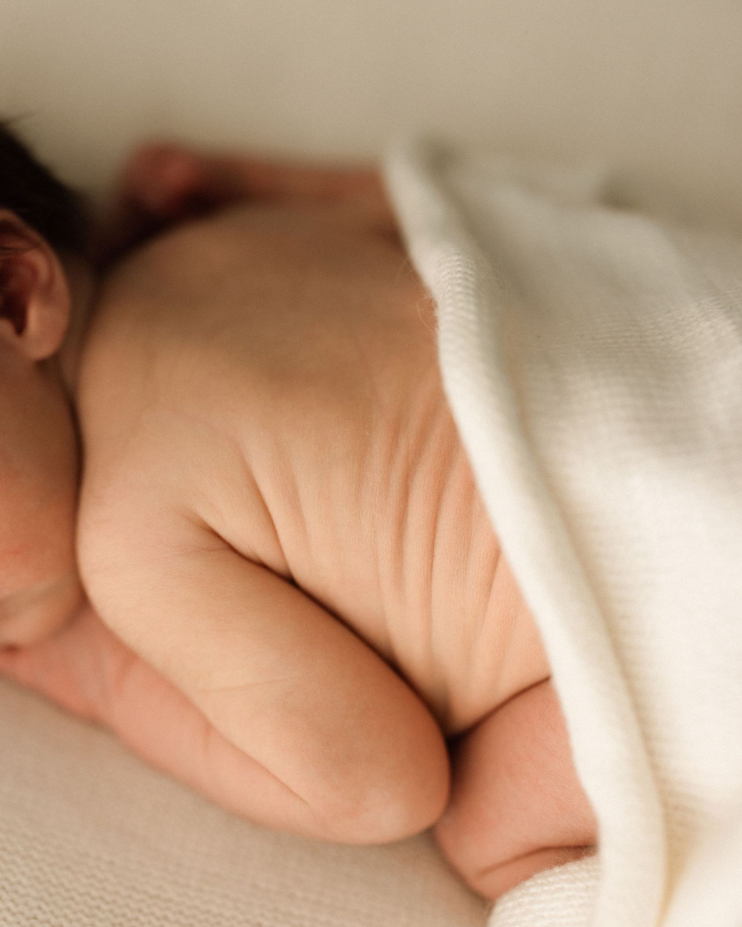 Newborn baby sleeping peacefully on side, wrapped in soft white knit blanket, showing chubby legs and back.