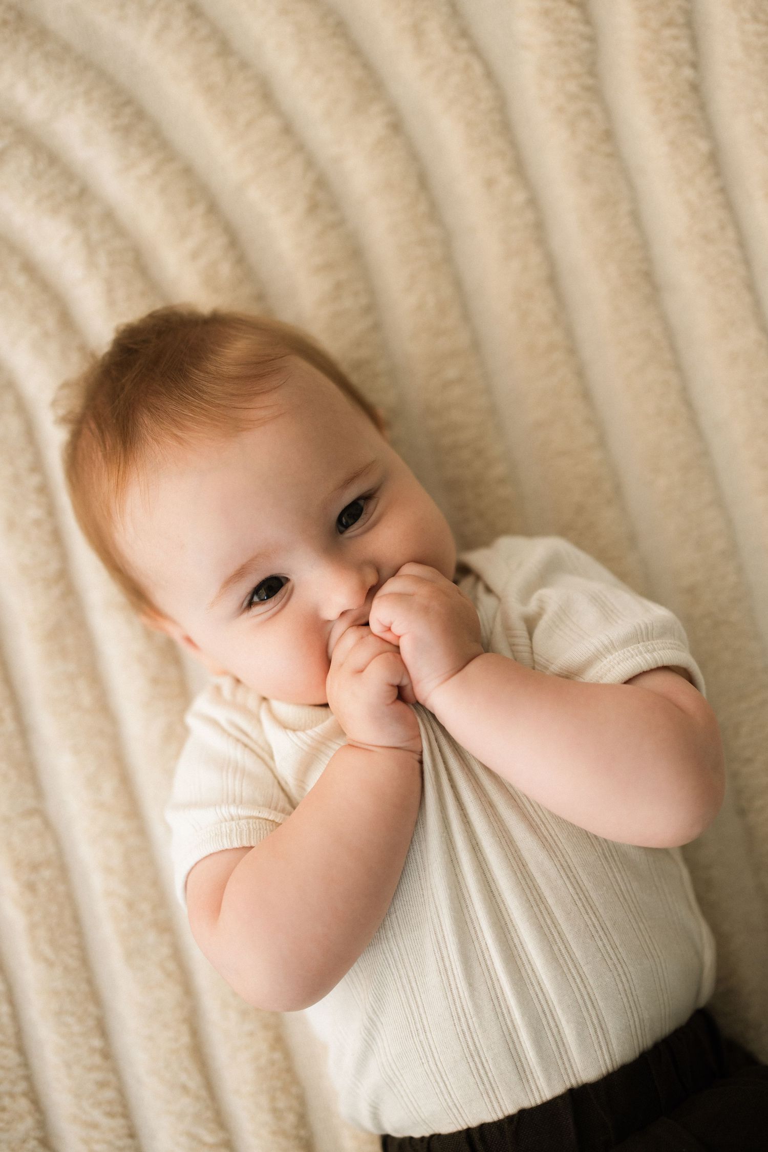Baby in cream sweater lying on textured white blanket while sucking thumb.