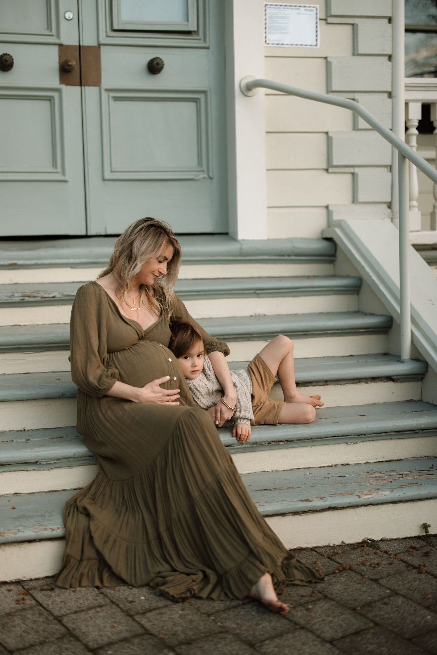 A pregnant person in a long brown dress sits with a small child on gray stone steps in front of a mint green door.