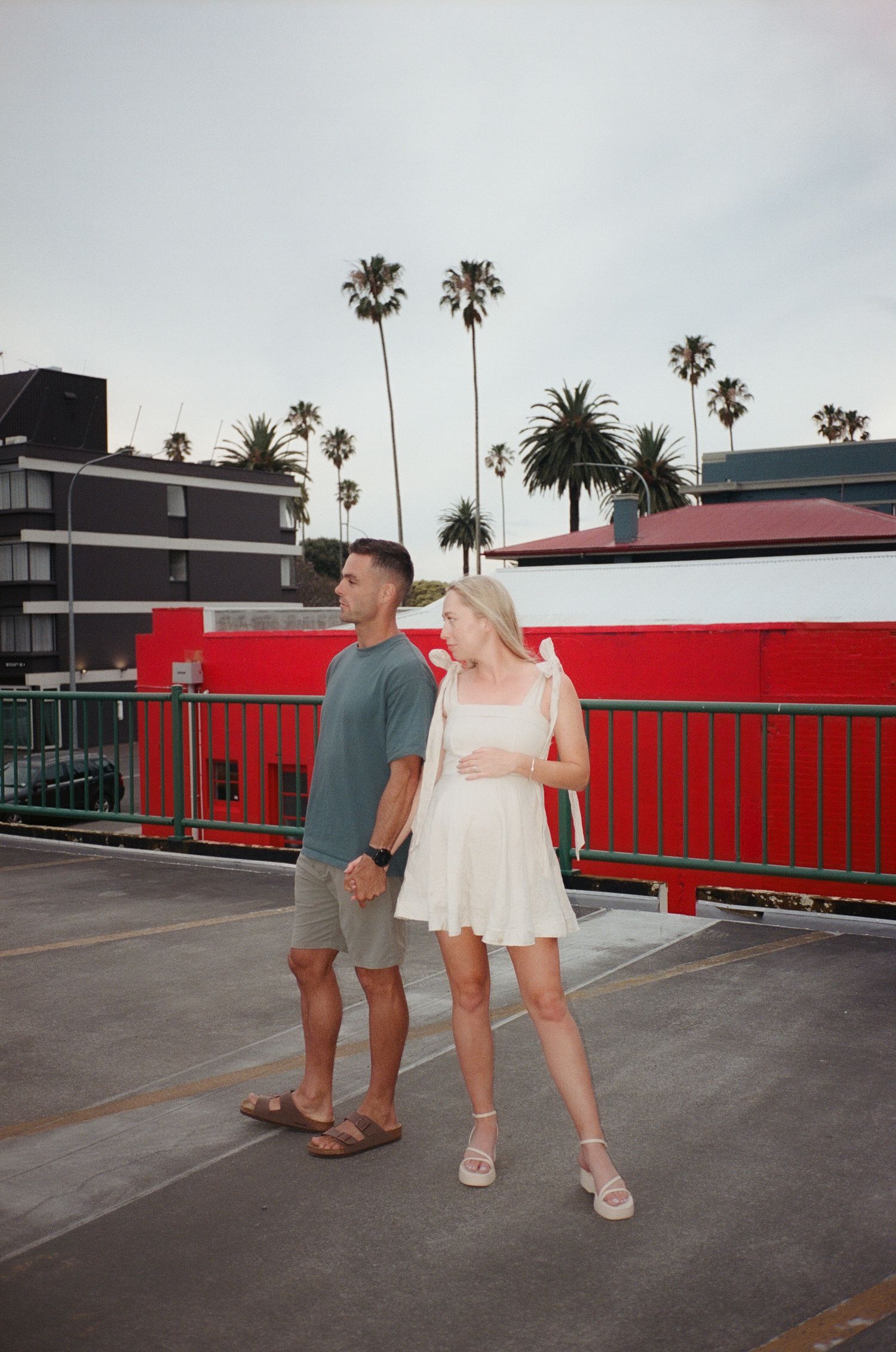 Couple holding hands on rooftop parking lot with palm trees and red building backdrop.
