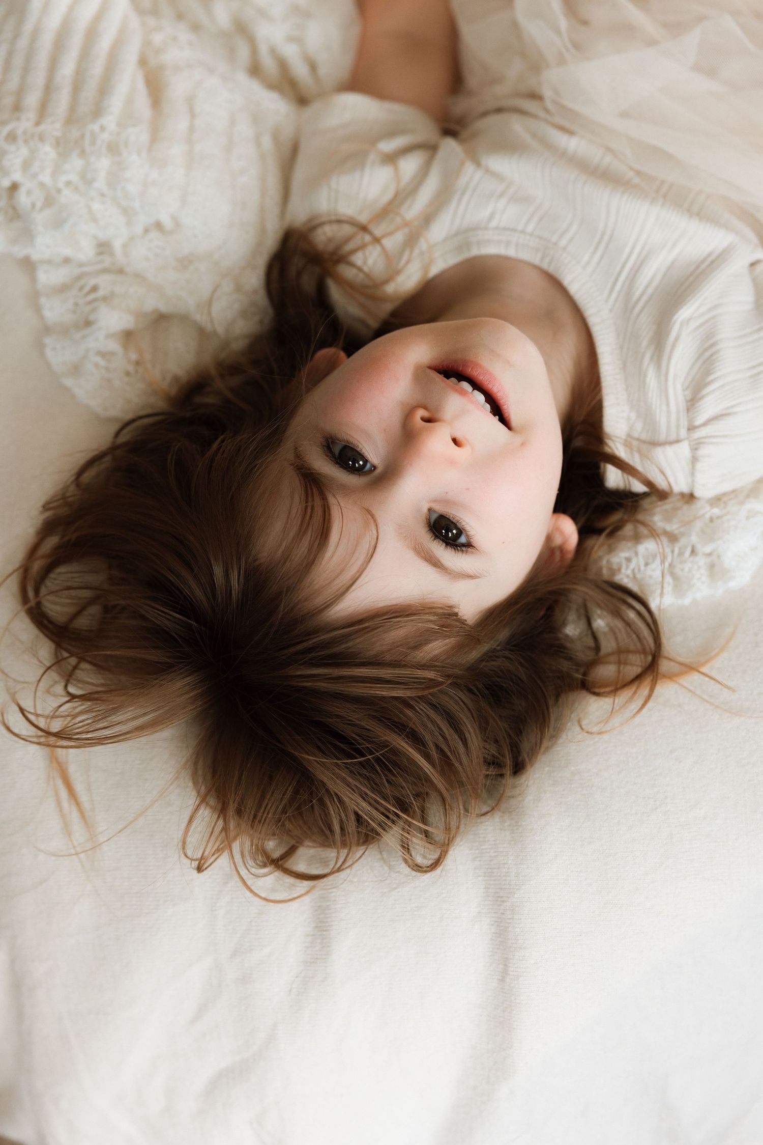 A young person with brown hair lies on white bedding while gazing upward at the camera.