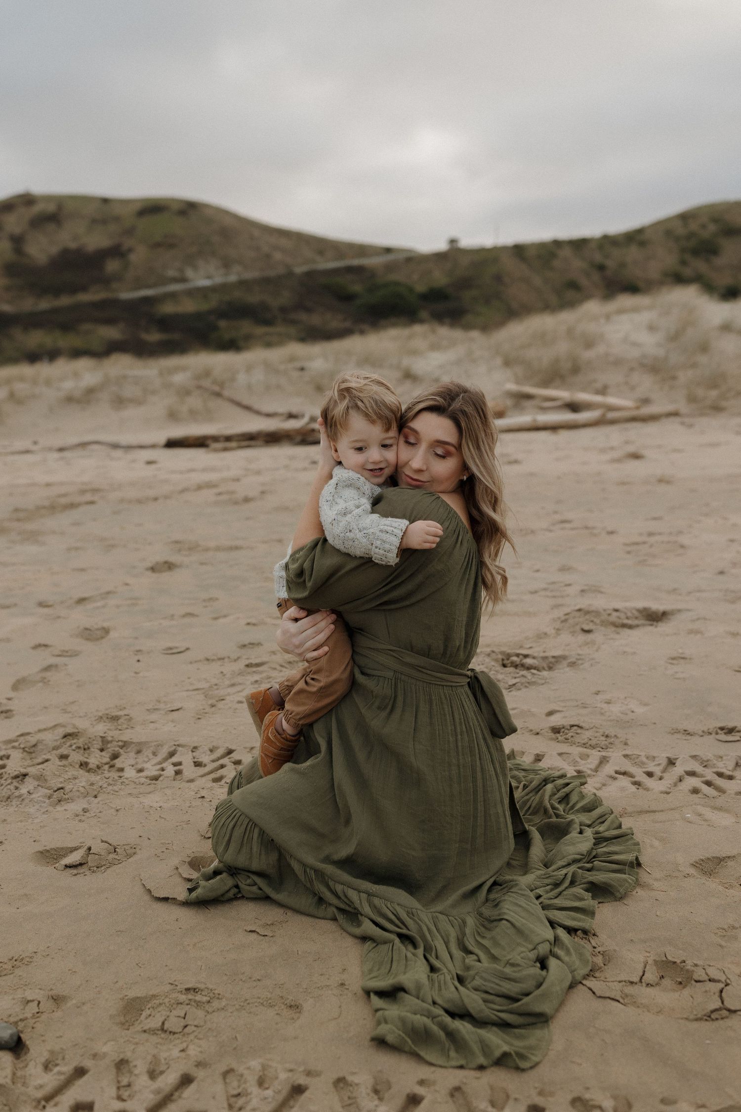 A tender moment captured on a sandy beach with flowing green dress and overcast sky.