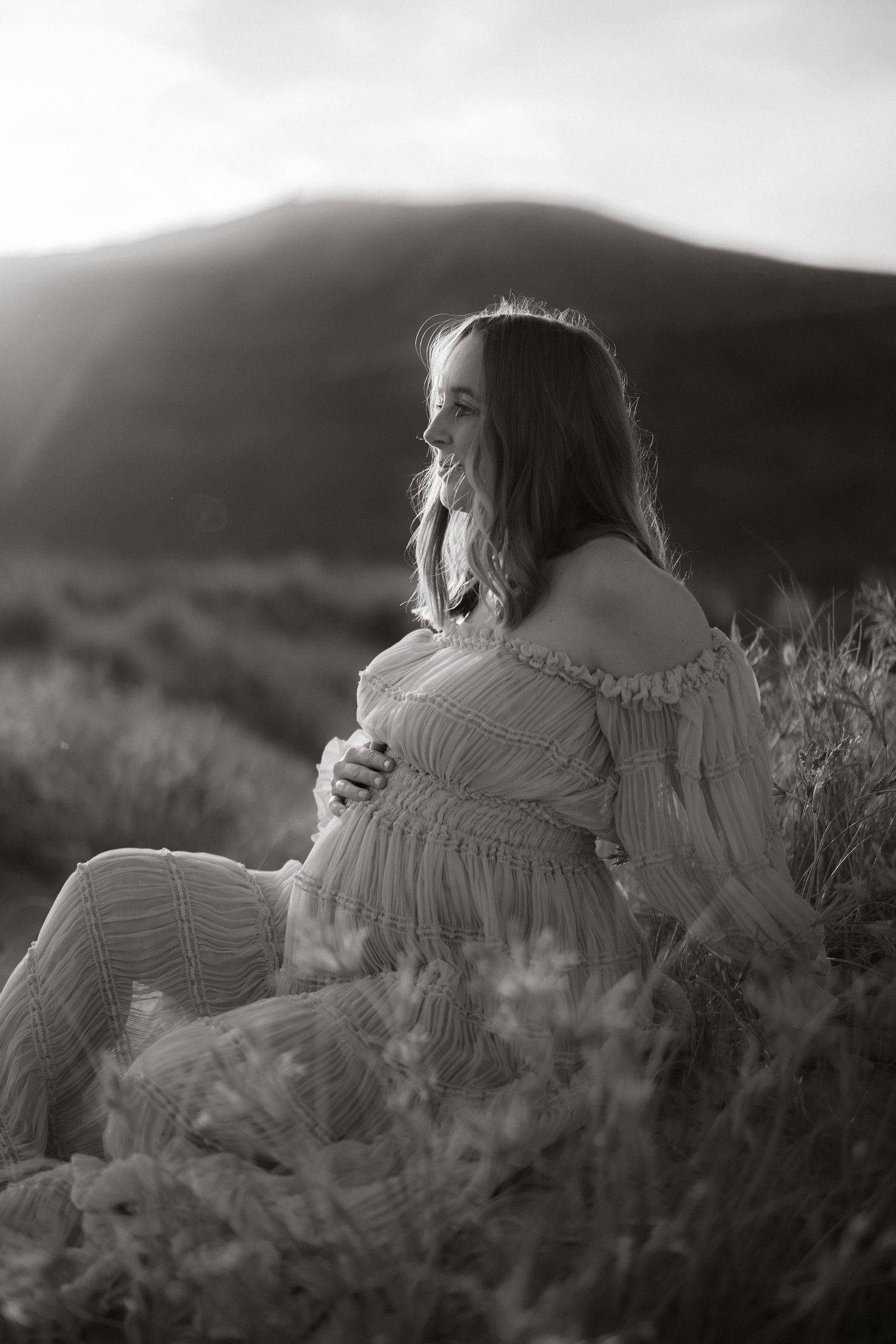 A scenic maternity portrait in black and white showing a woman sitting among wildflowers at sunset.