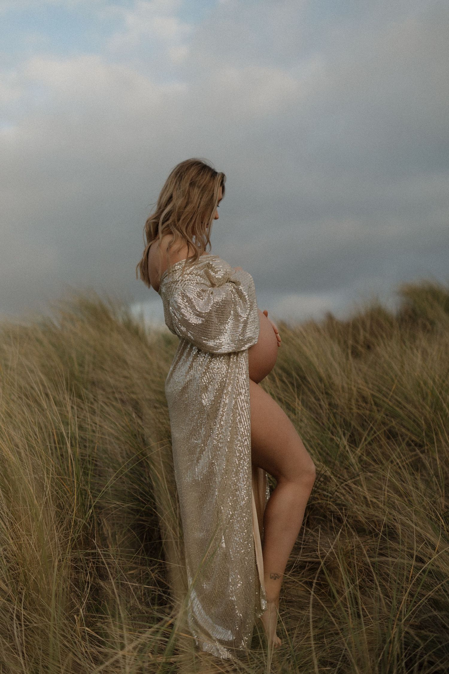 Figure in sparkly dress standing among windswept beach grass at dusk.