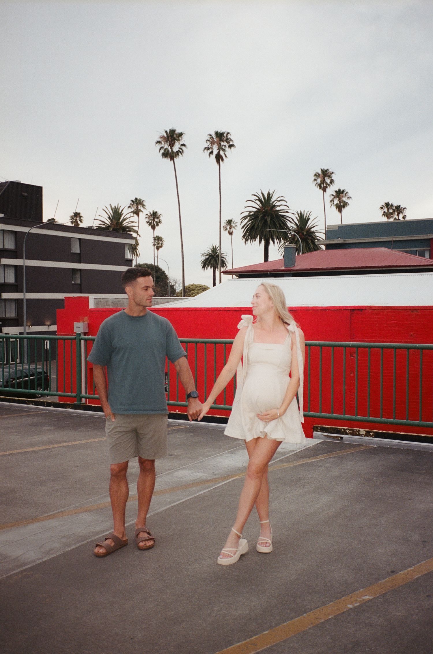 Couple holding hands on rooftop parking lot with palm trees and red building in sunny California backdrop.