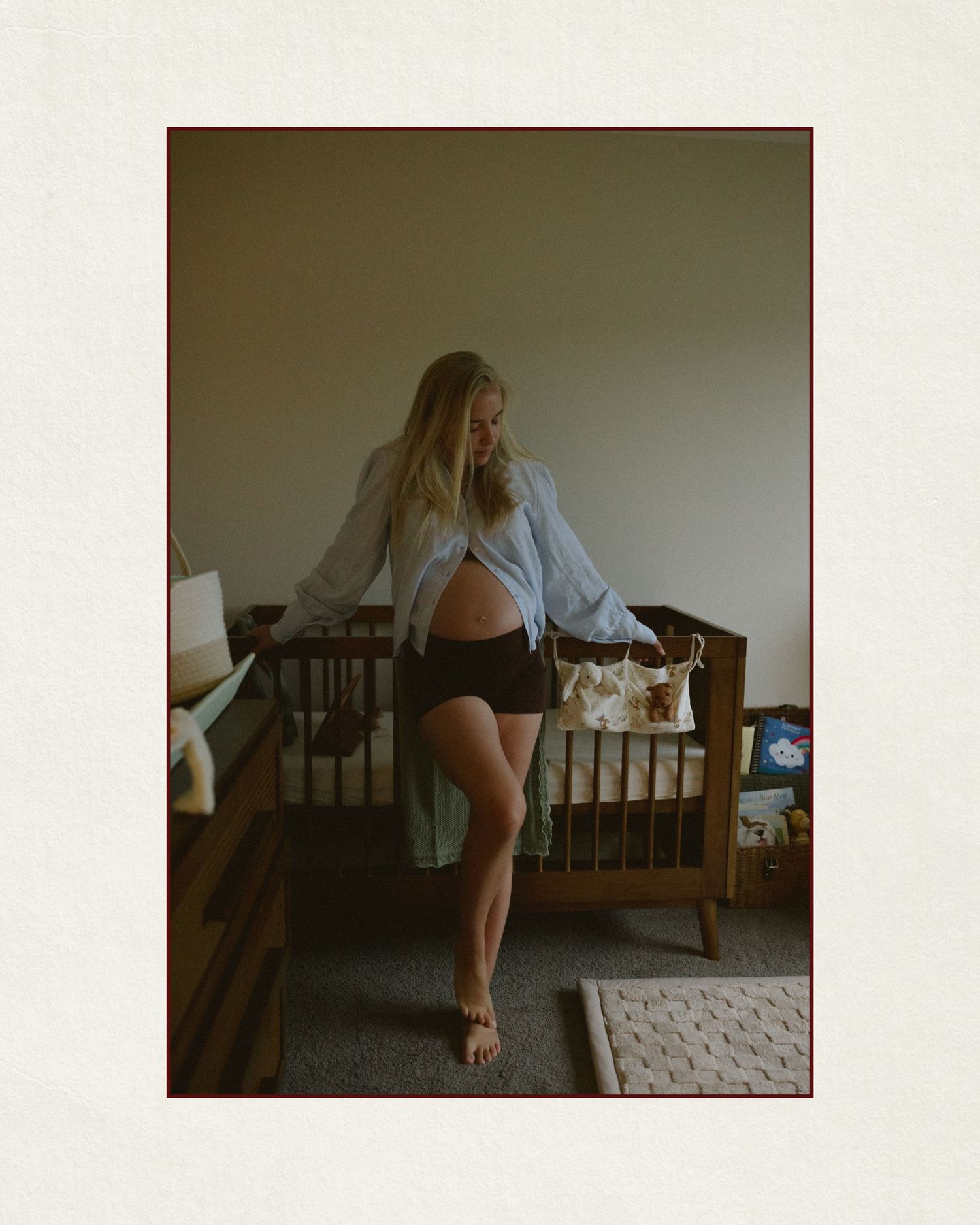 Pregnant woman in blue shirt and black shorts standing in nursery room with wooden crib.