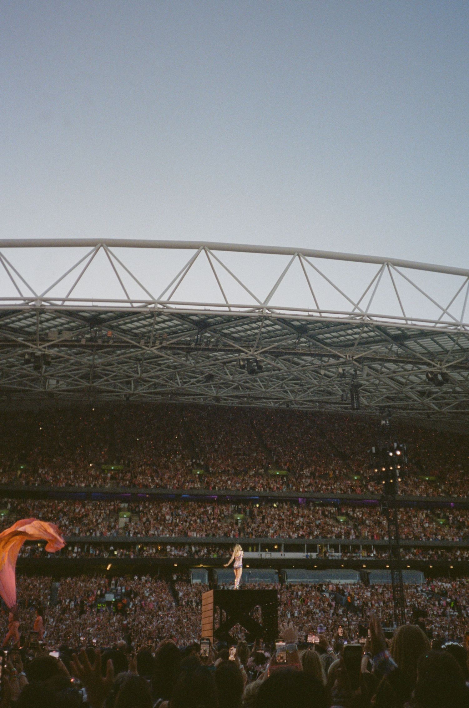Massive stadium concert with packed crowd, performer on stage, structural roof visible against clear sky.