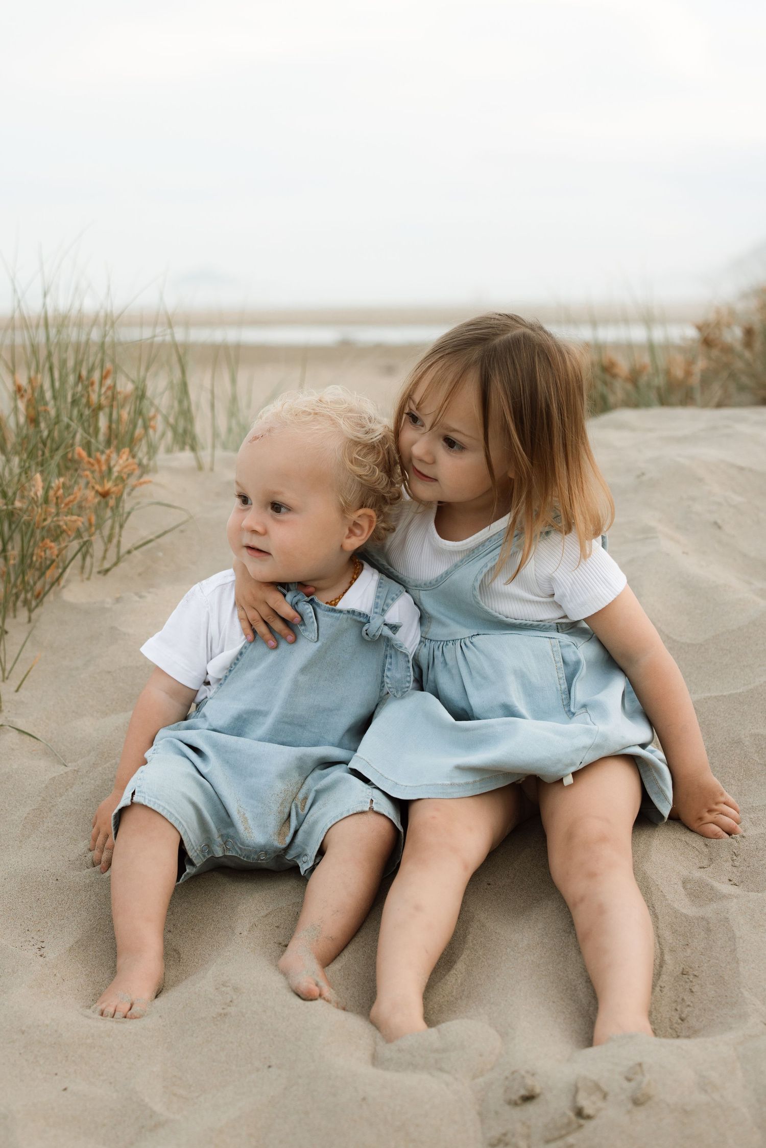 Two children in light blue outfits sitting together on sandy beach during overcast day.