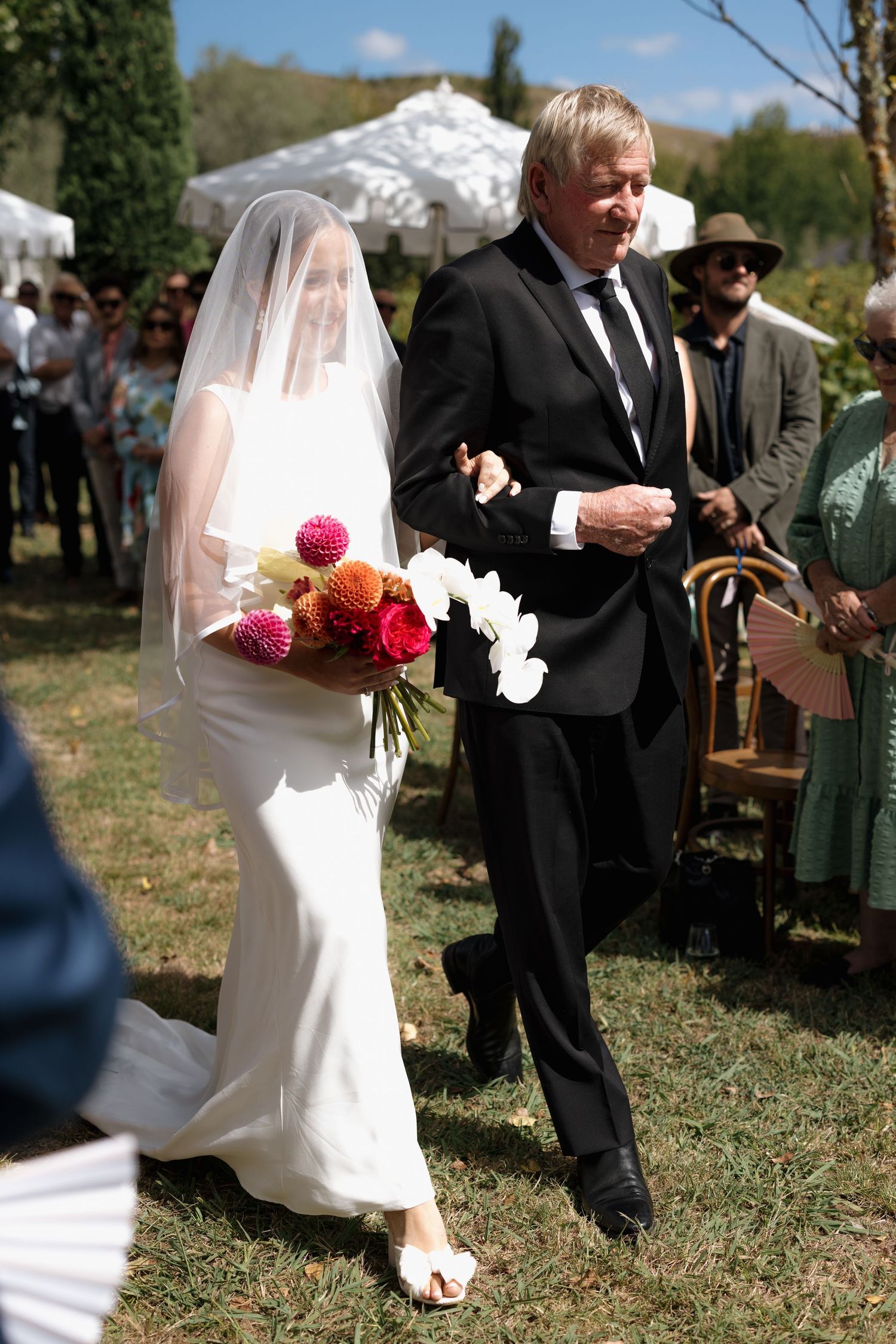 A bride in white holds a colorful bouquet while walking down an outdoor wedding aisle with an escort.