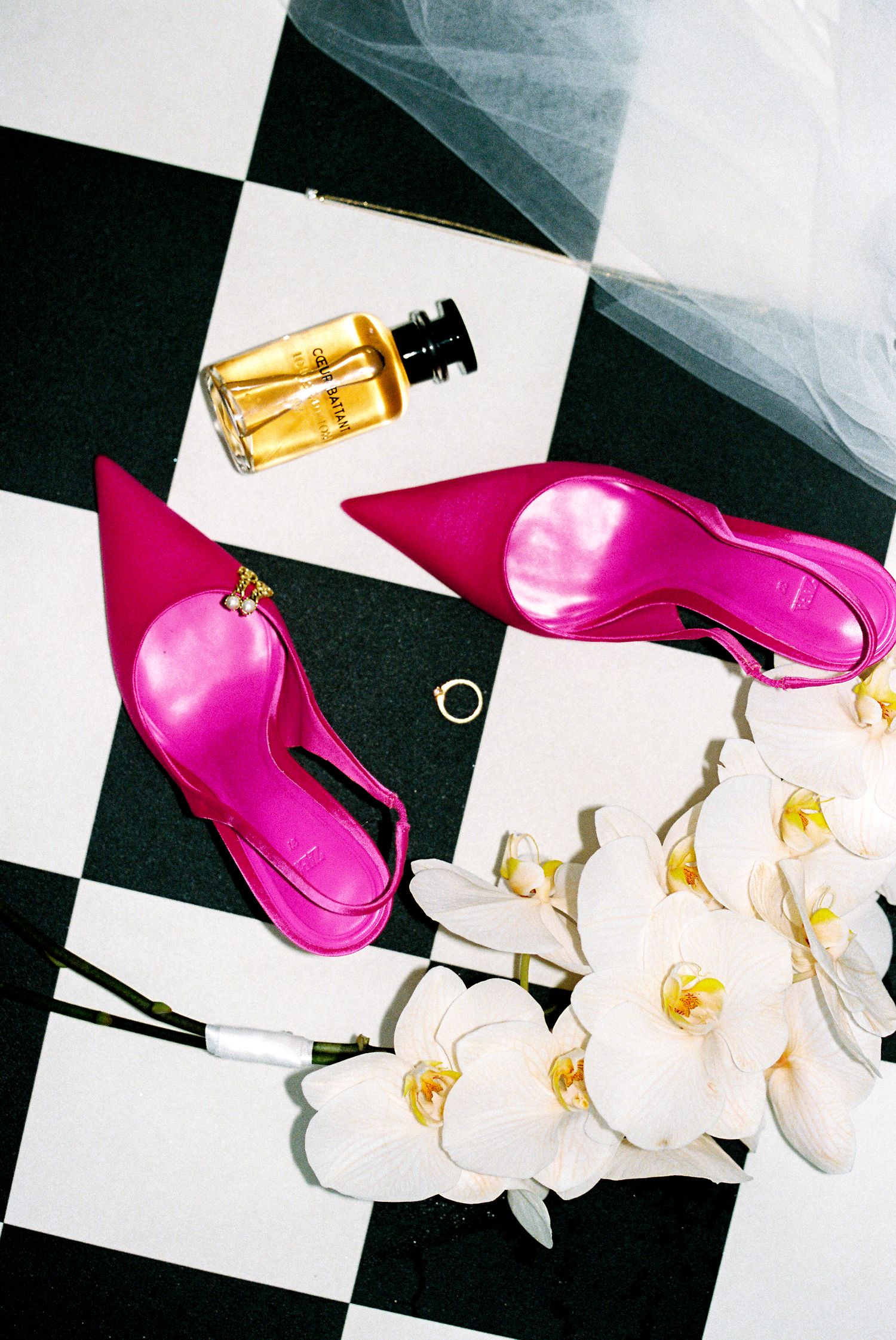 Pink heels, perfume bottle, and white orchids arranged on black and white checkered floor.