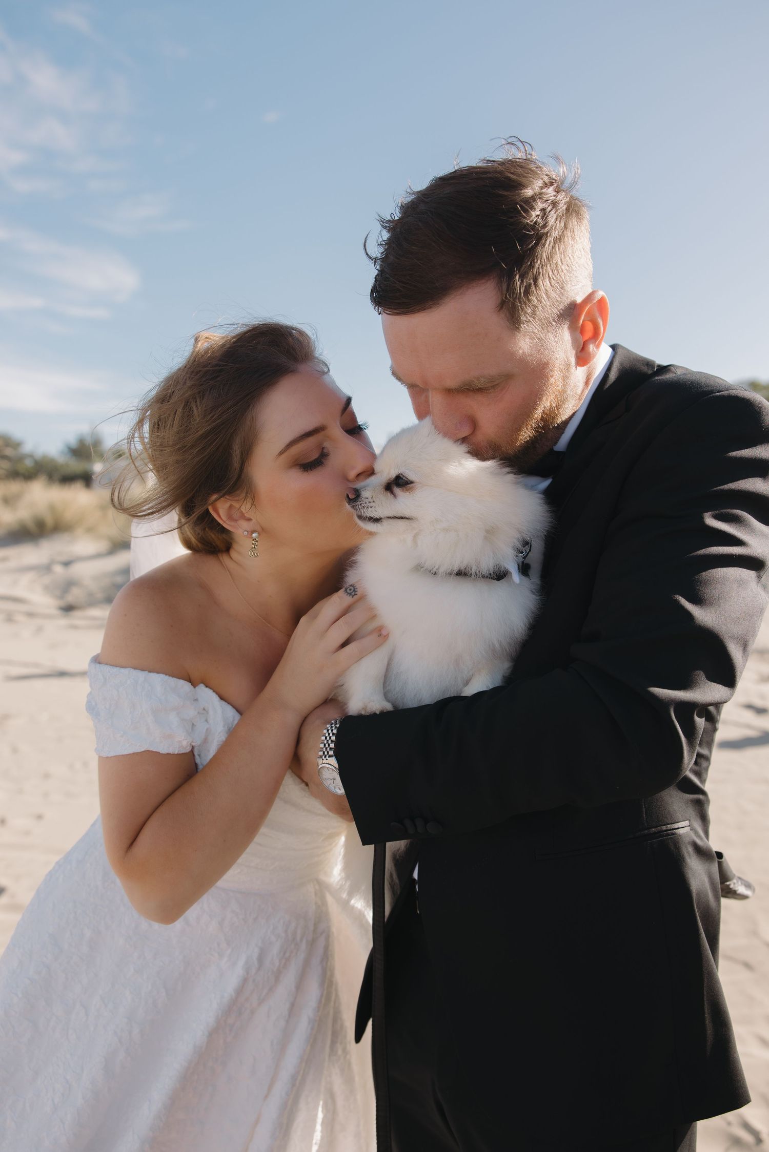 A couple shares an intimate moment kissing their fluffy white dog in desert sunlight.