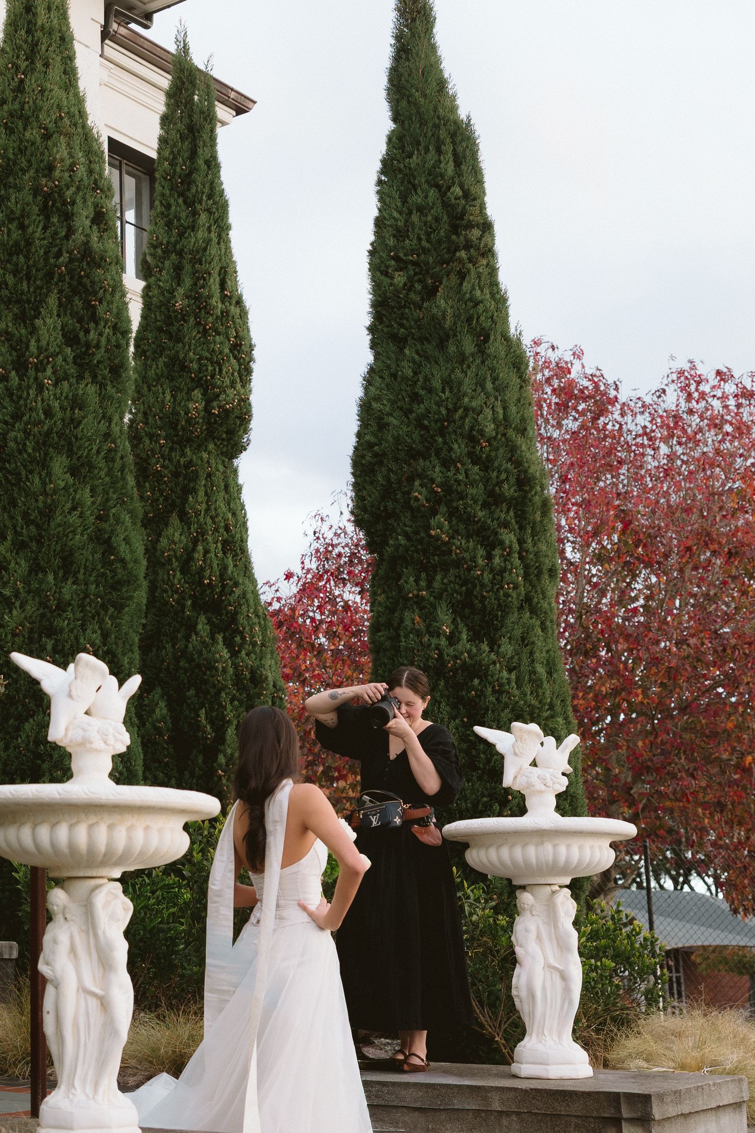 Bride in white gown and veil stands near ornate white fountains as bridesmaid adjusts her hair outdoors.