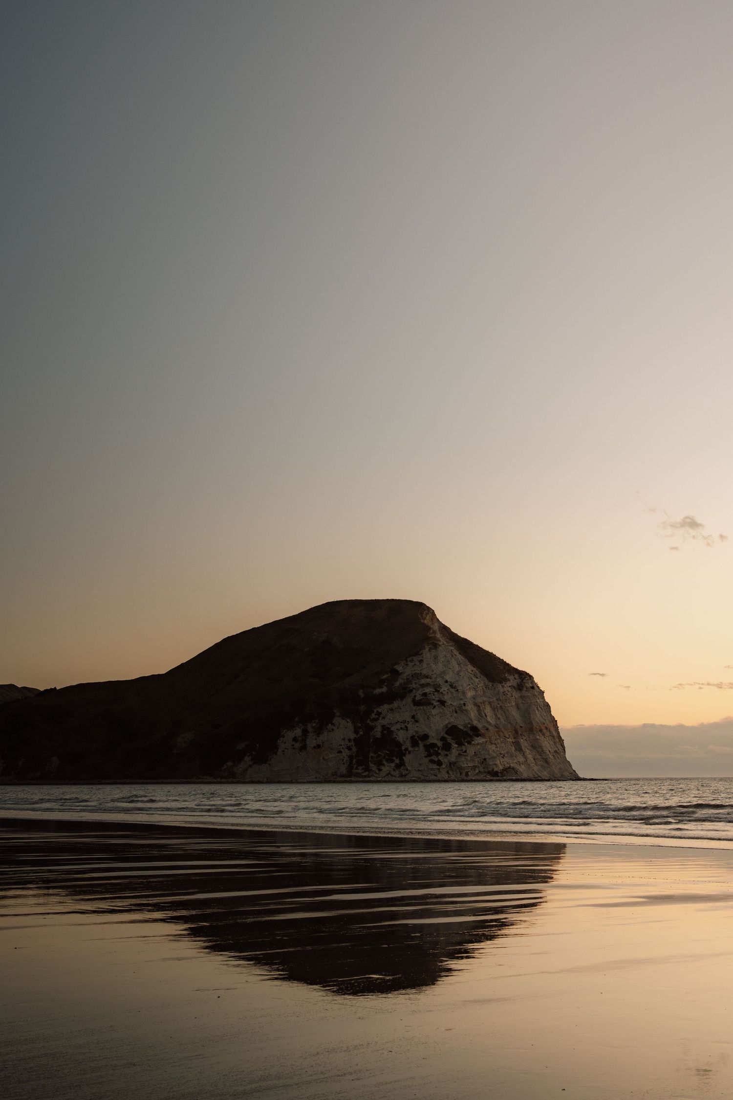 Dramatic coastal sunset with a silhouetted chalk headland reflected on wet sand, calm ocean waters, and a soft golden sky.