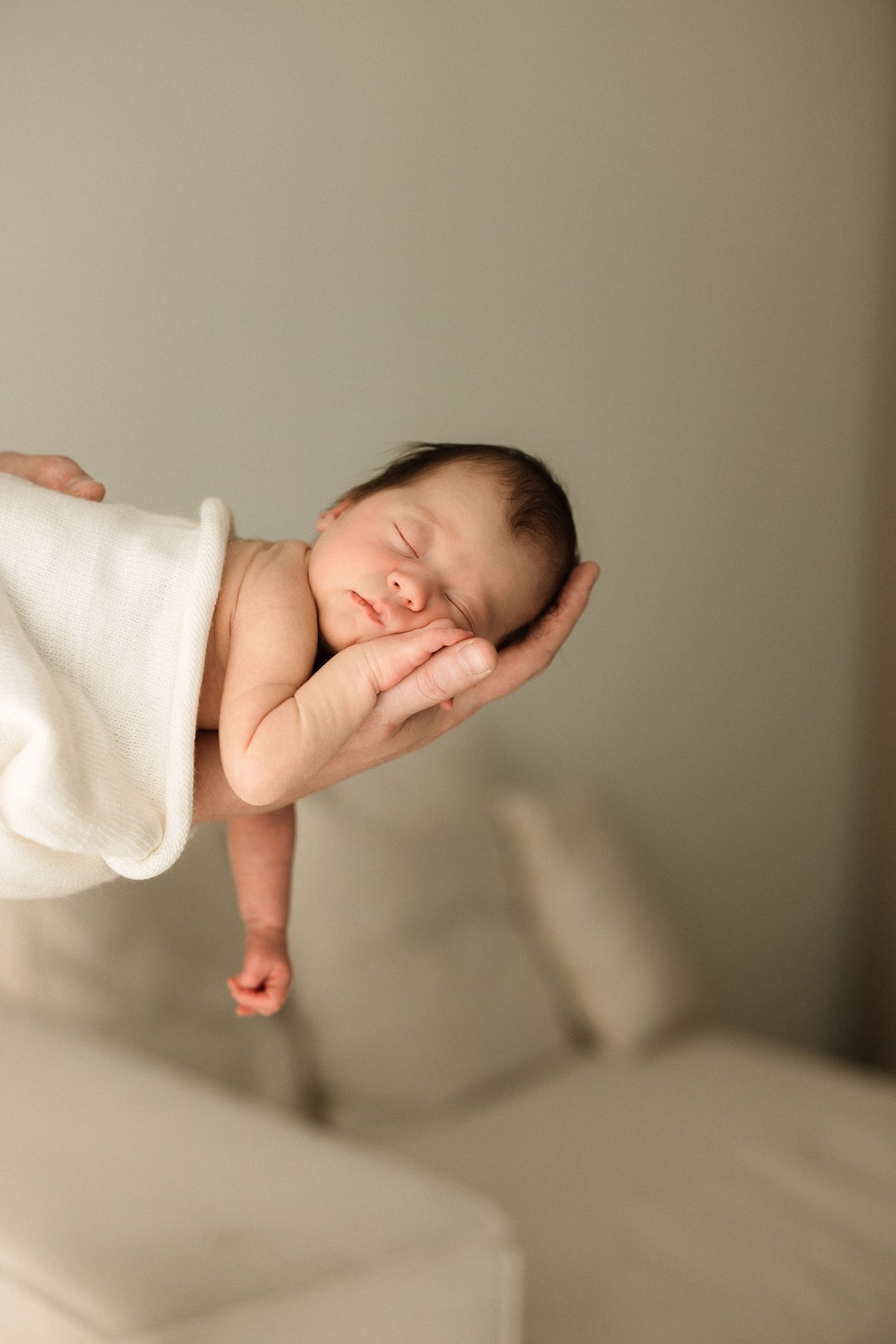 Sleeping newborn baby cradled in adult hands, wrapped in white blanket, peaceful infant portrait photography.