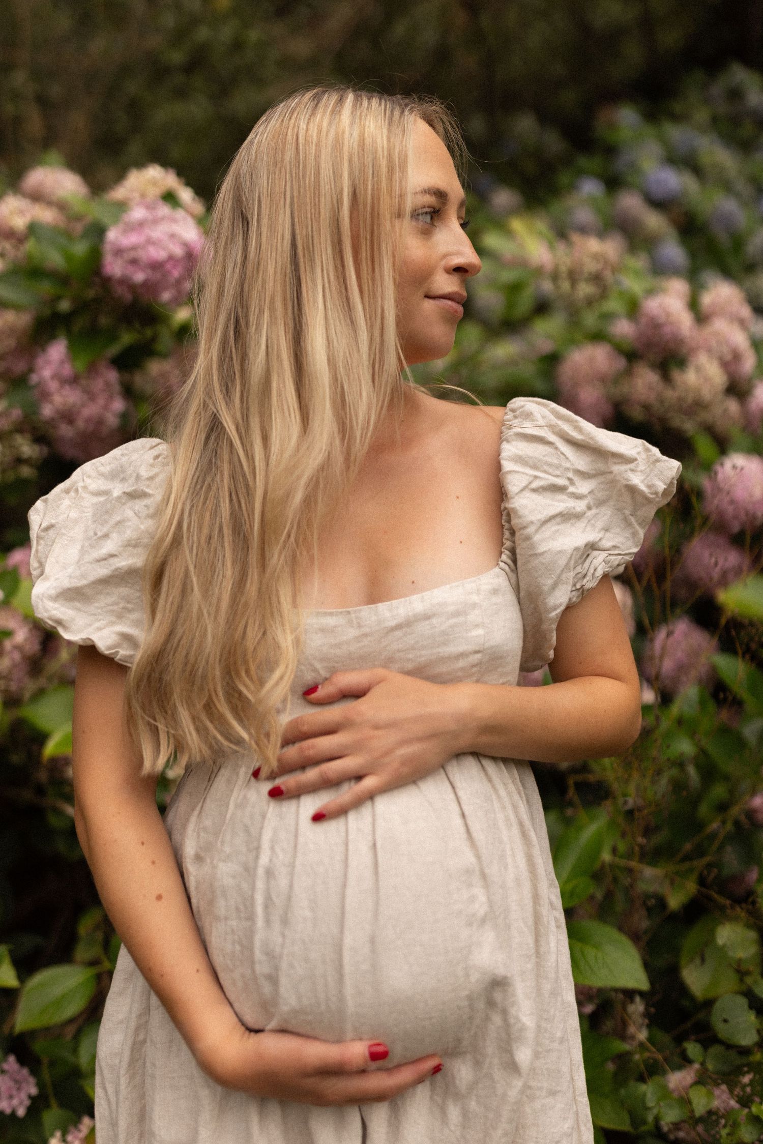 Pregnant woman in white dress cradling baby bump outdoors among blooming pink and purple hydrangeas.